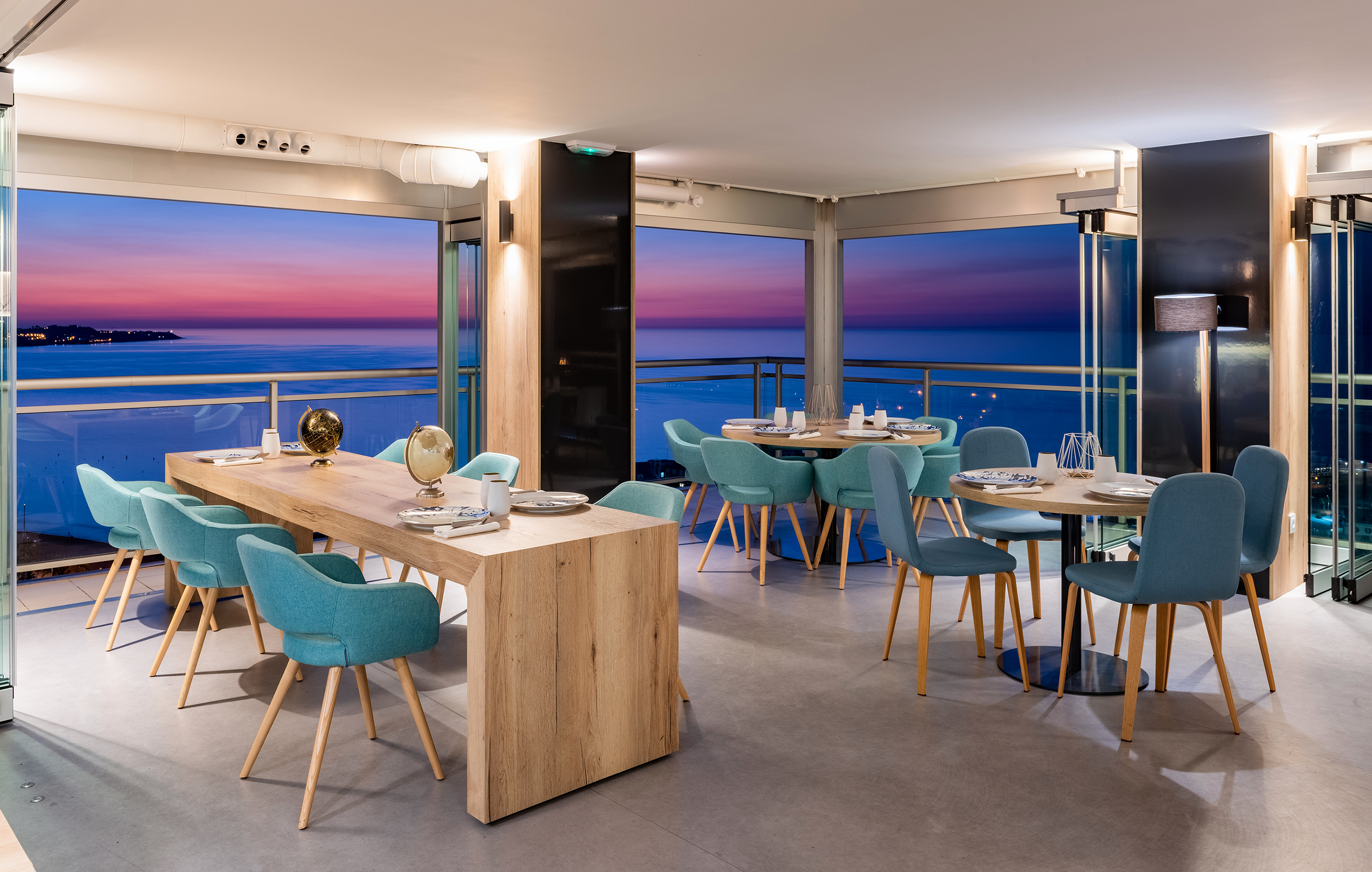 Modern restaurant dining room with ocean view at sunset, teal chairs, wooden tables.