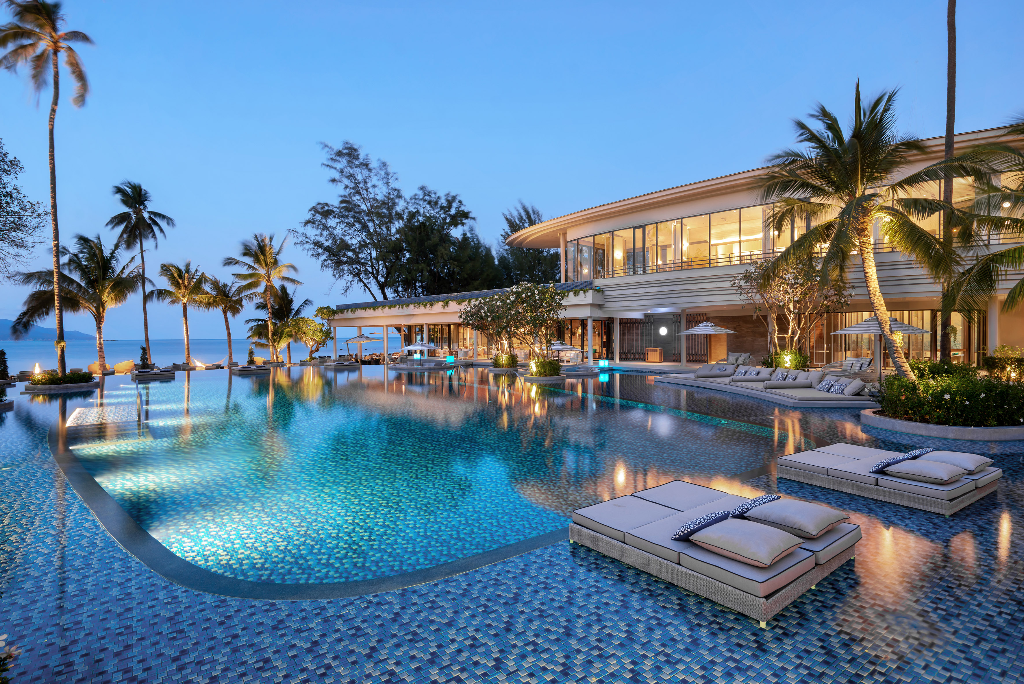 Luxury tropical resort with illuminated infinity pool, palm trees, ocean view.