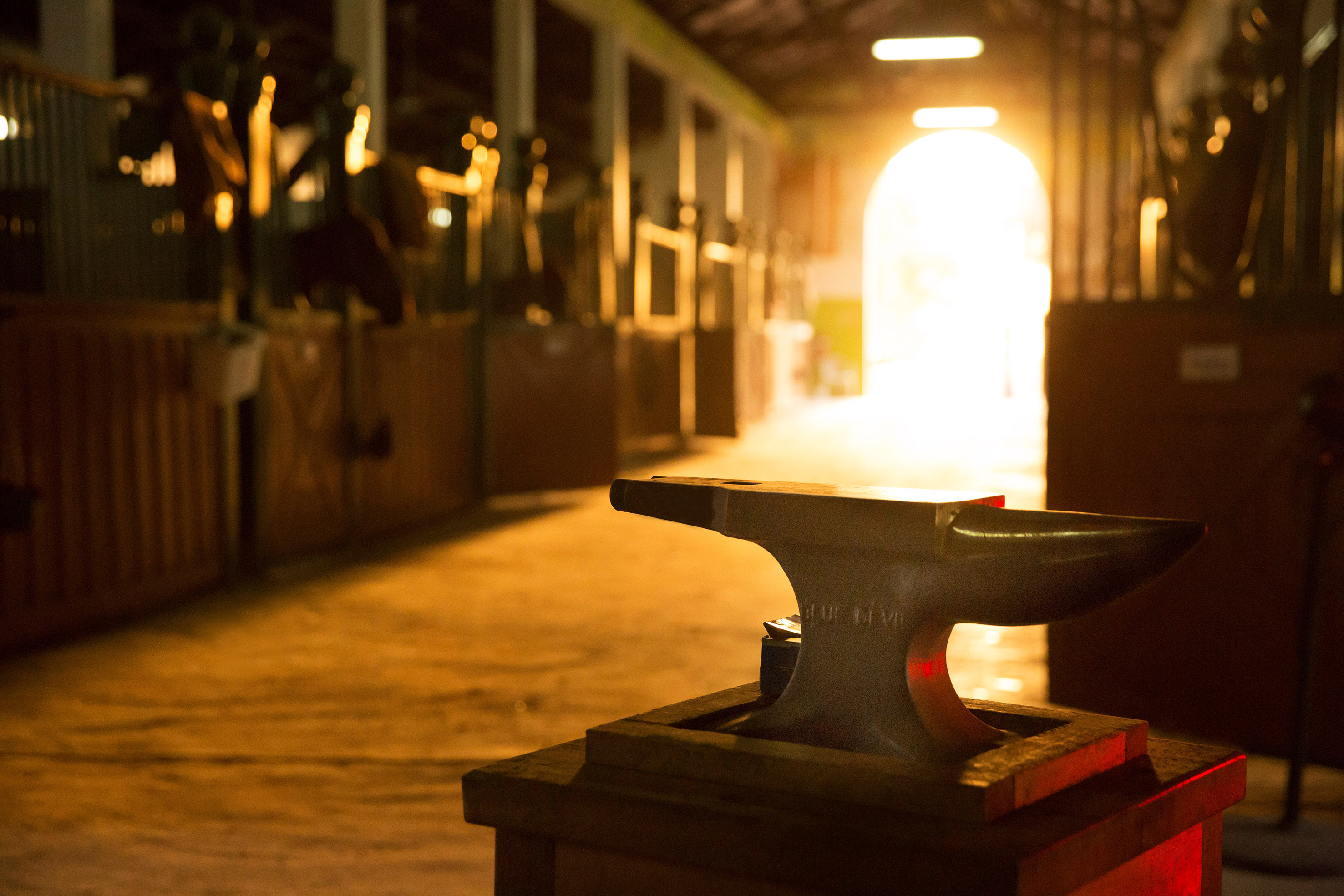Blacksmith's anvil in a warmly lit barn aisle with horse stalls.