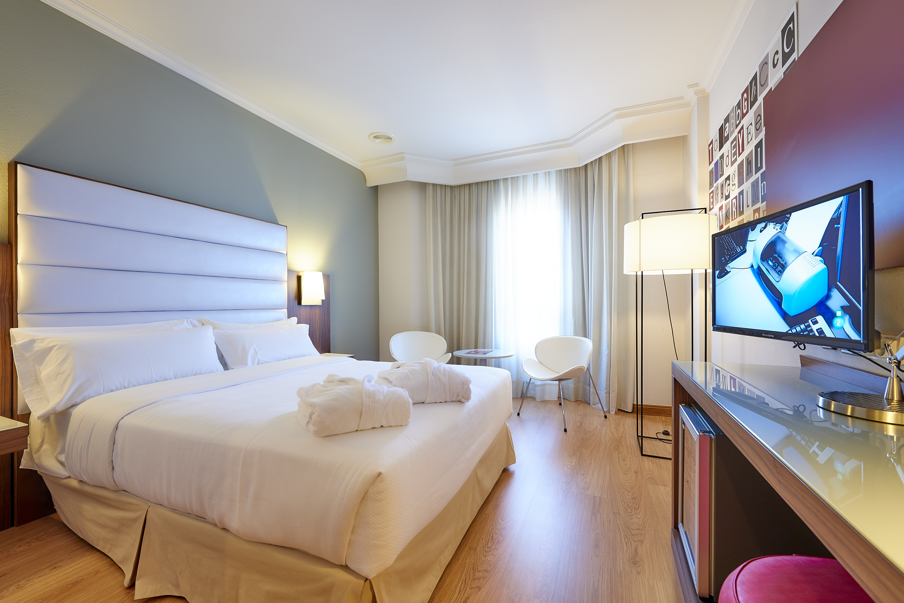 Modern hotel room with large bed, white bathrobes, TV, and bright window.