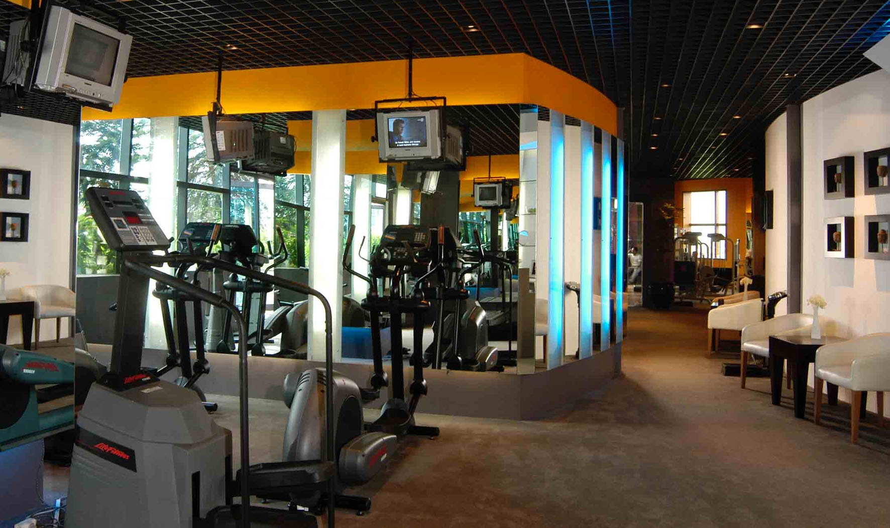 Modern fitness center with cardio equipment, ceiling TVs, mirrored walls, and lounge seating.