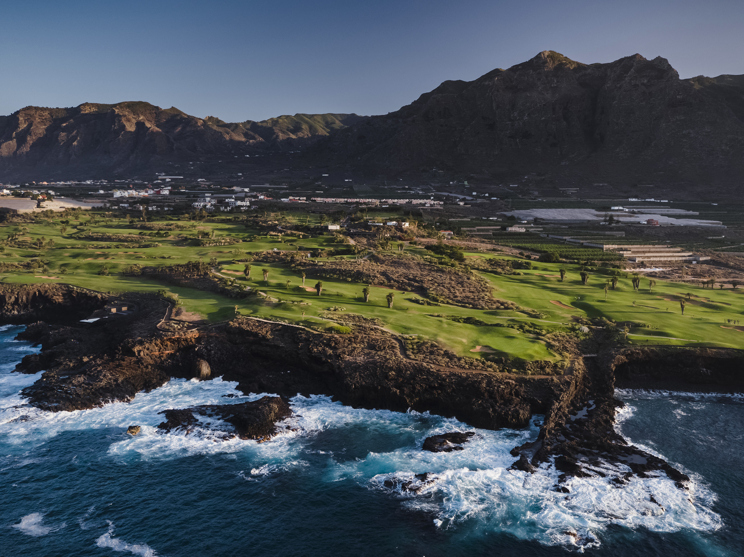 Green coastal golf course with ocean waves, volcanic cliffs, and mountains.