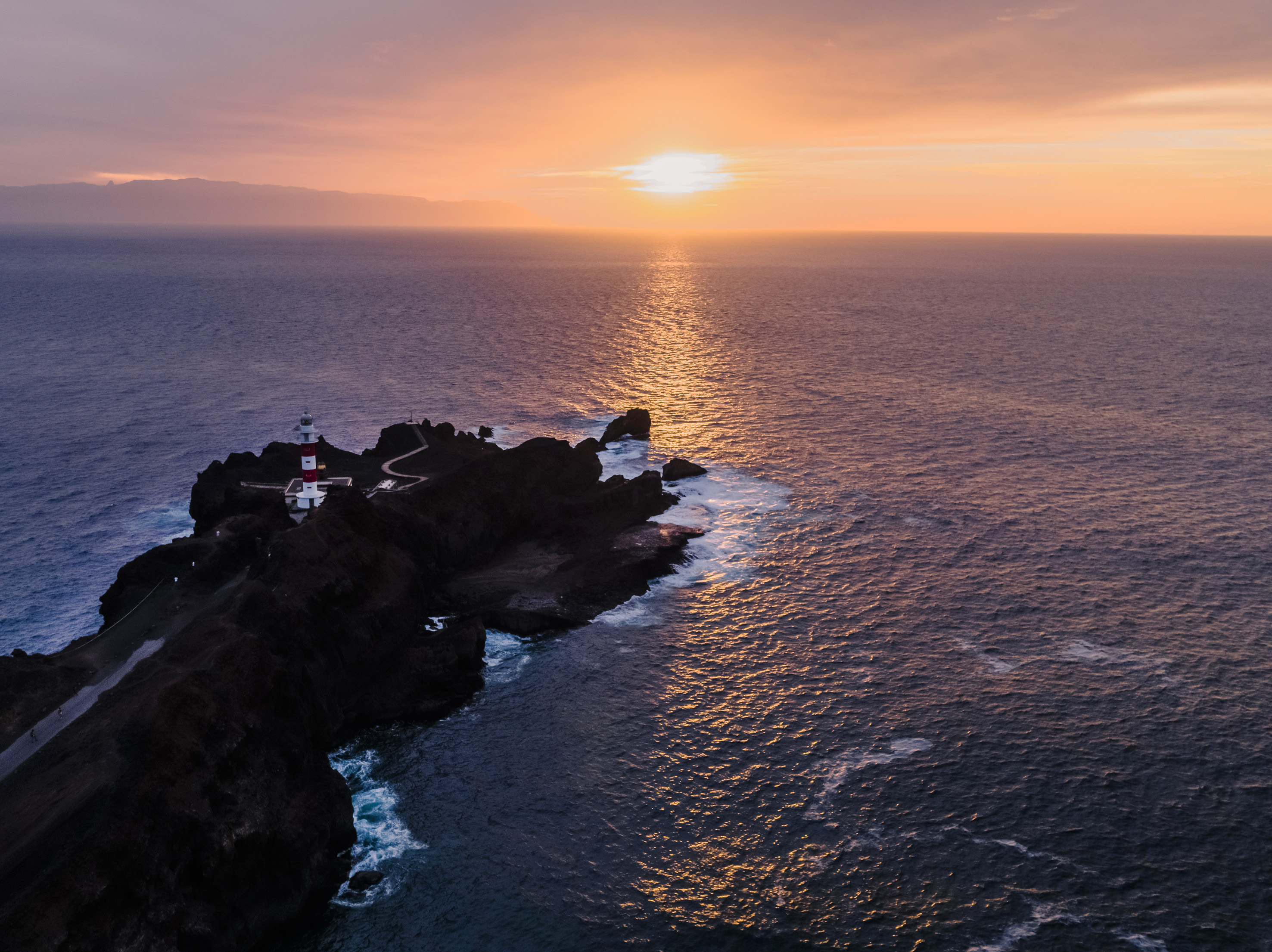 Aerial view: Lighthouse on rocky peninsula, vibrant sunset over the ocean.