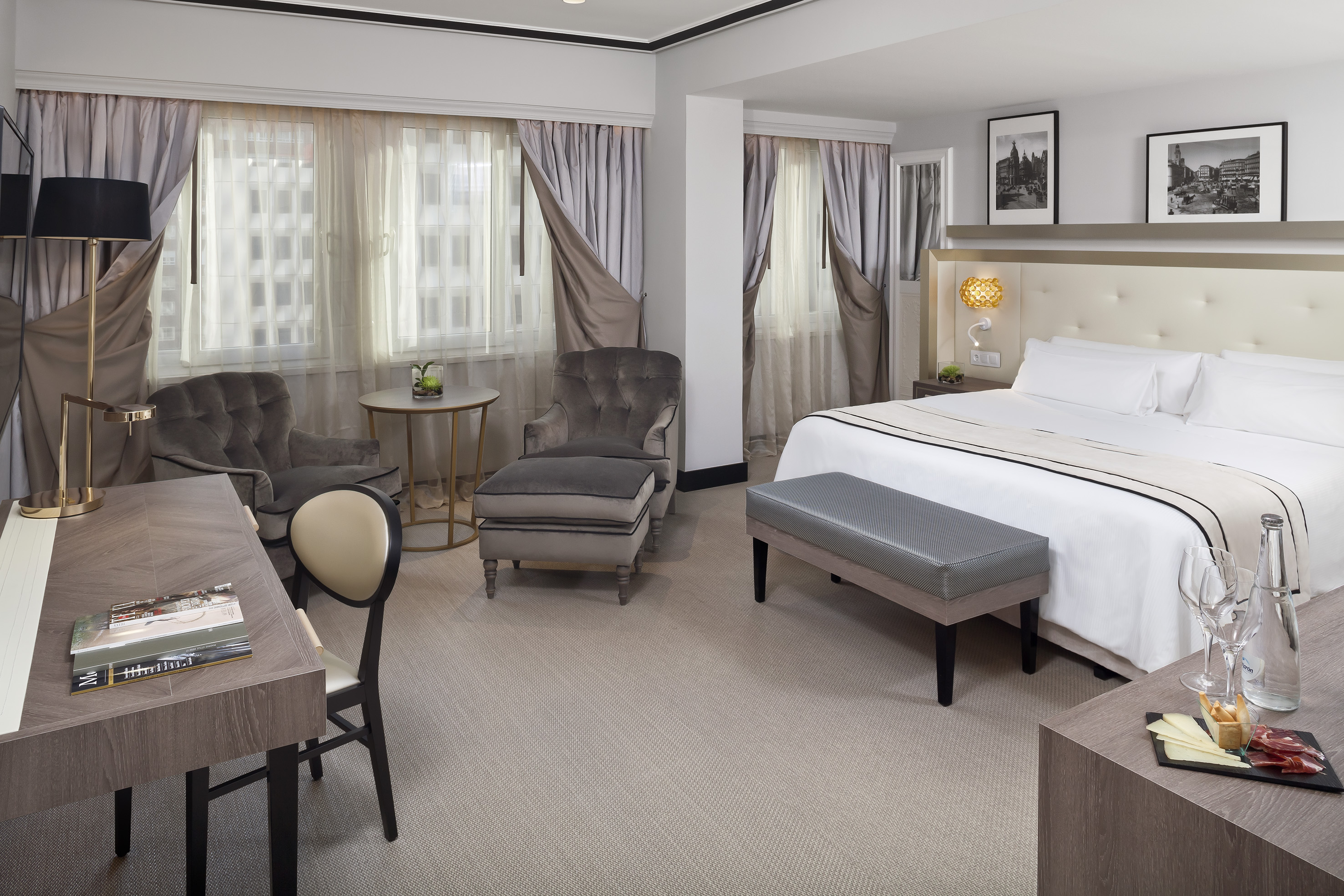 Luxury hotel room with king bed, sitting area, desk, and large windows.