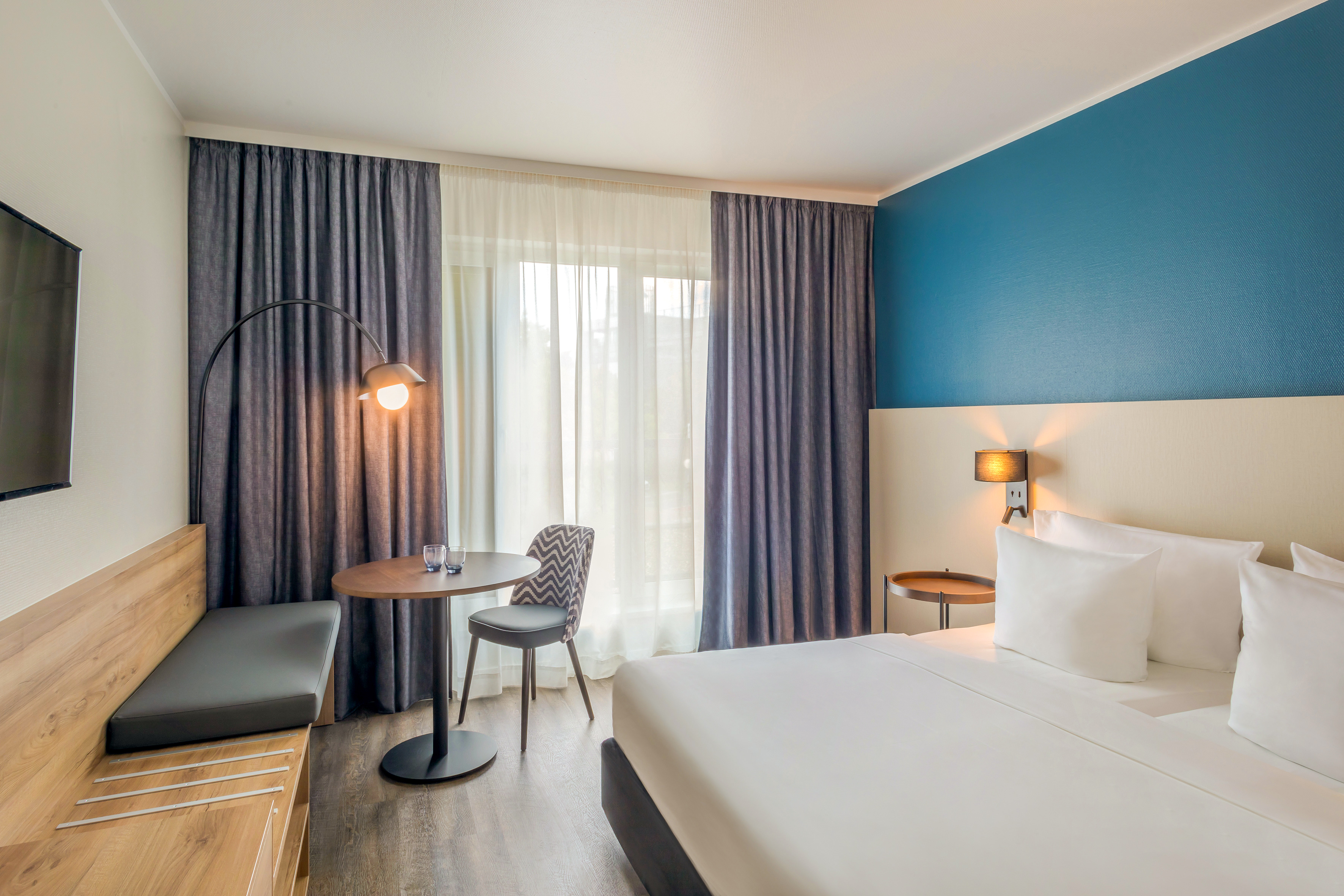 Modern hotel room with blue accent wall, bed, desk, and large window.