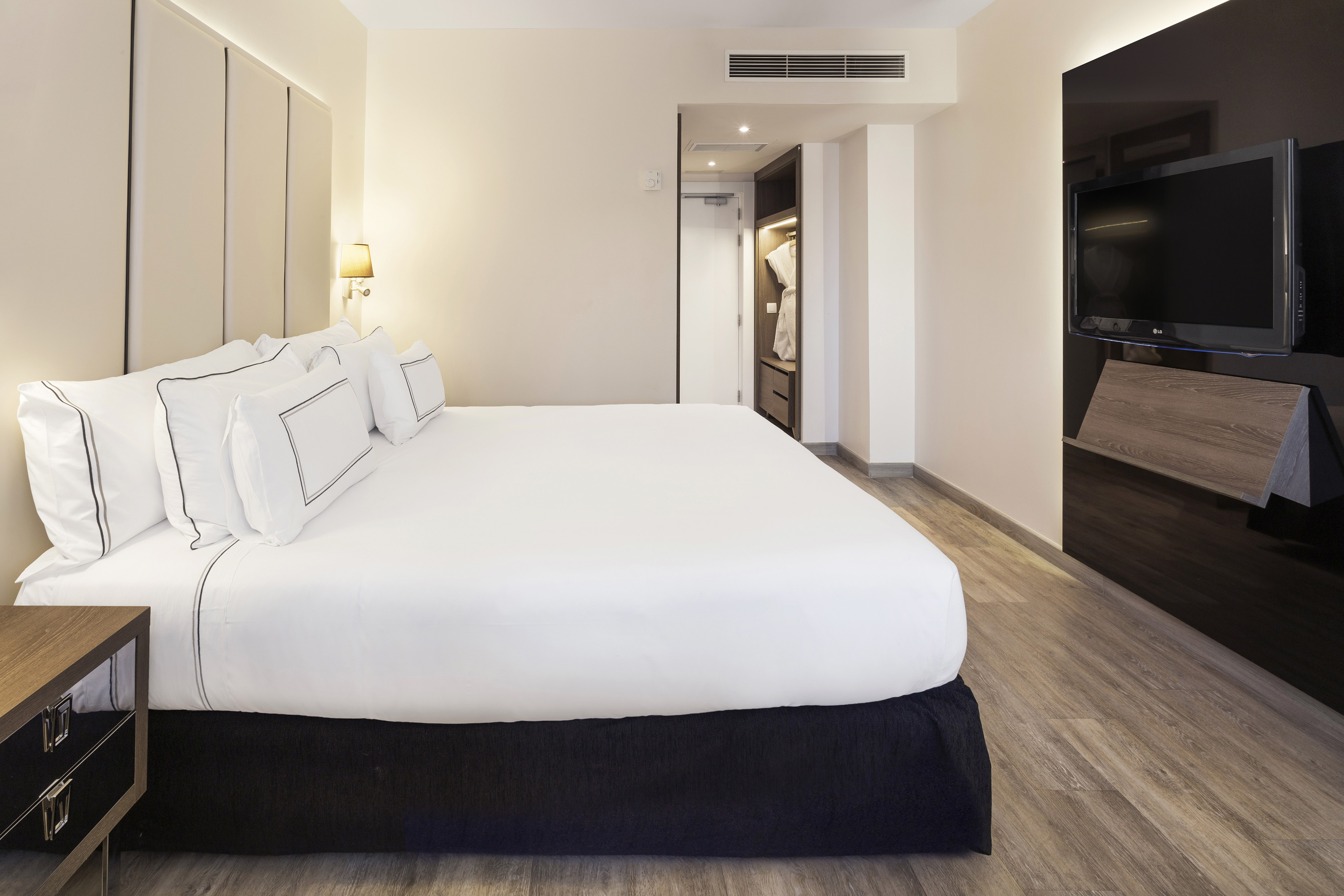 Hotel room with king bed, white sheets, cushioned headboard, and wall-mounted TV.