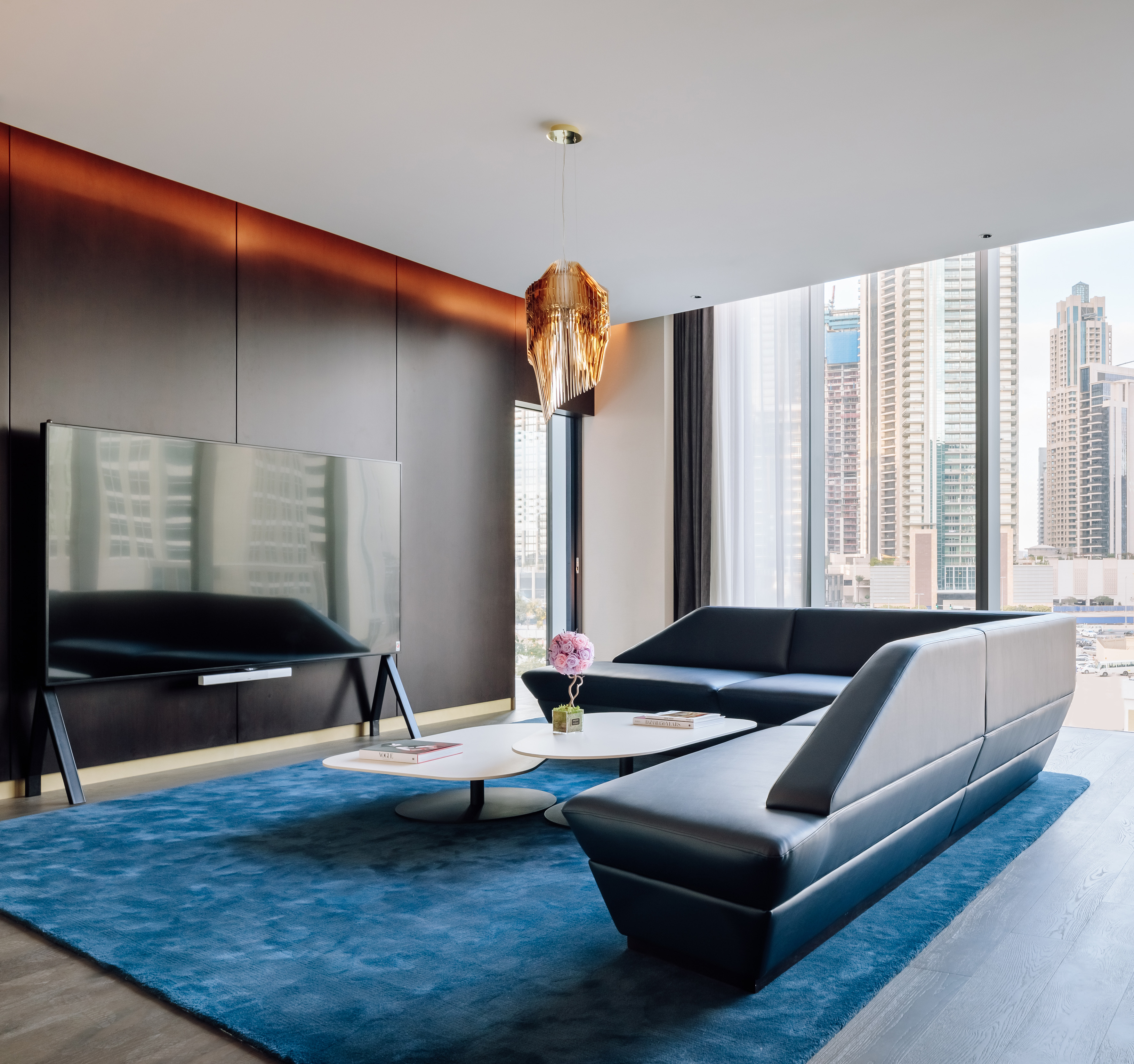 Modern living room with black sofas, blue rug, gold chandelier, and city view.