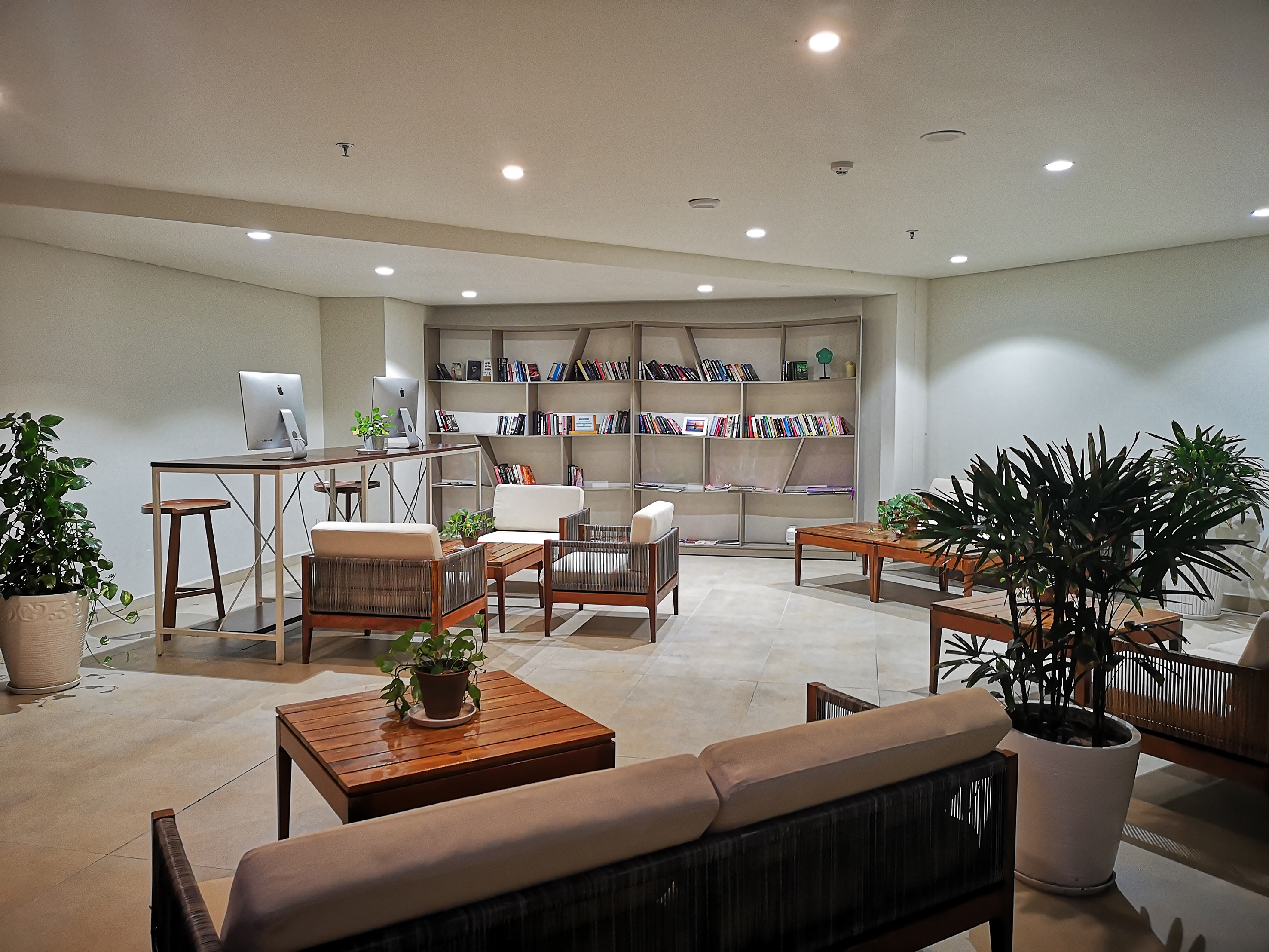 Modern lounge with iMacs, library bookshelves, comfortable seating, and potted plants.