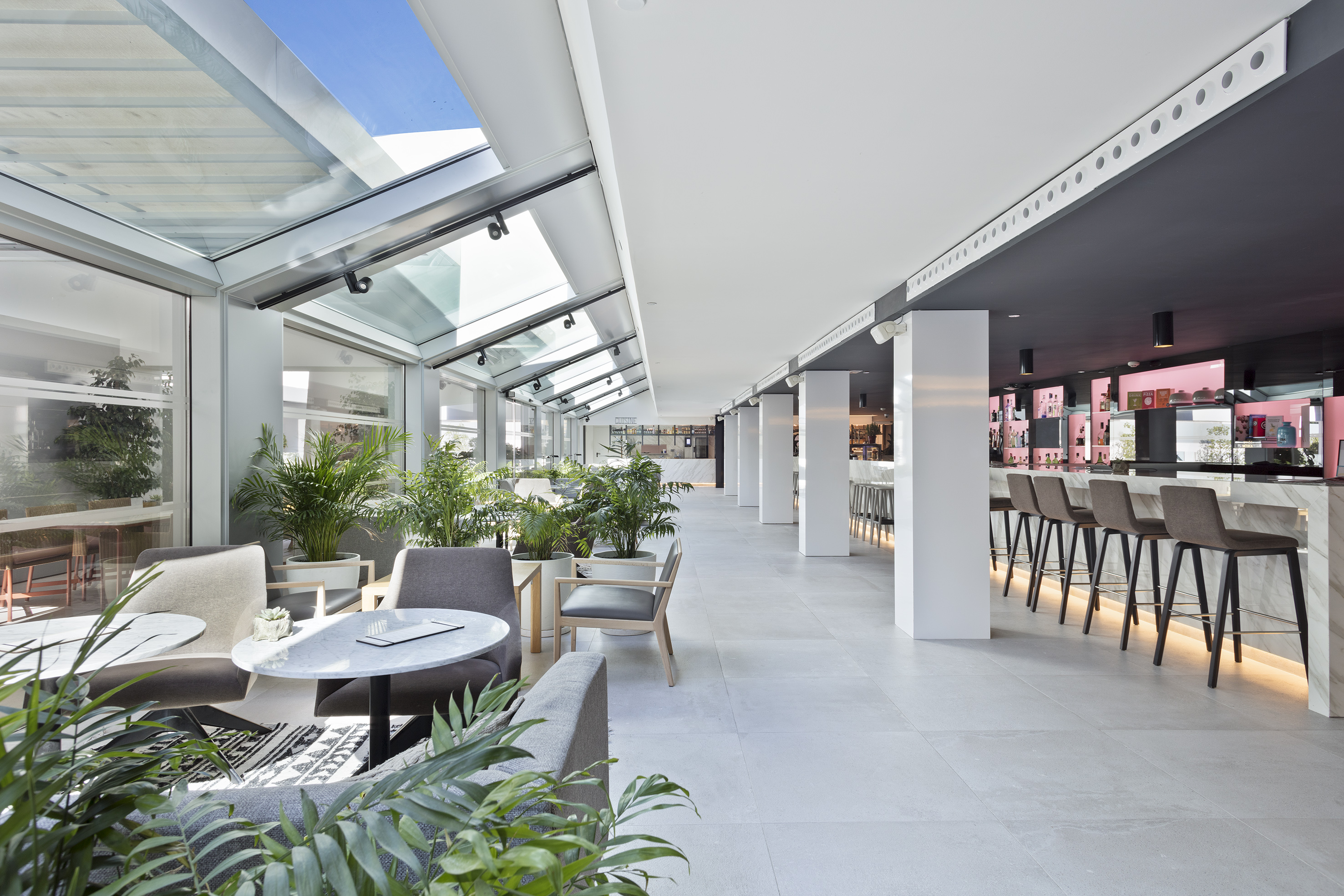 Modern lounge bar with glass roof, indoor plants, and sleek marble bar.