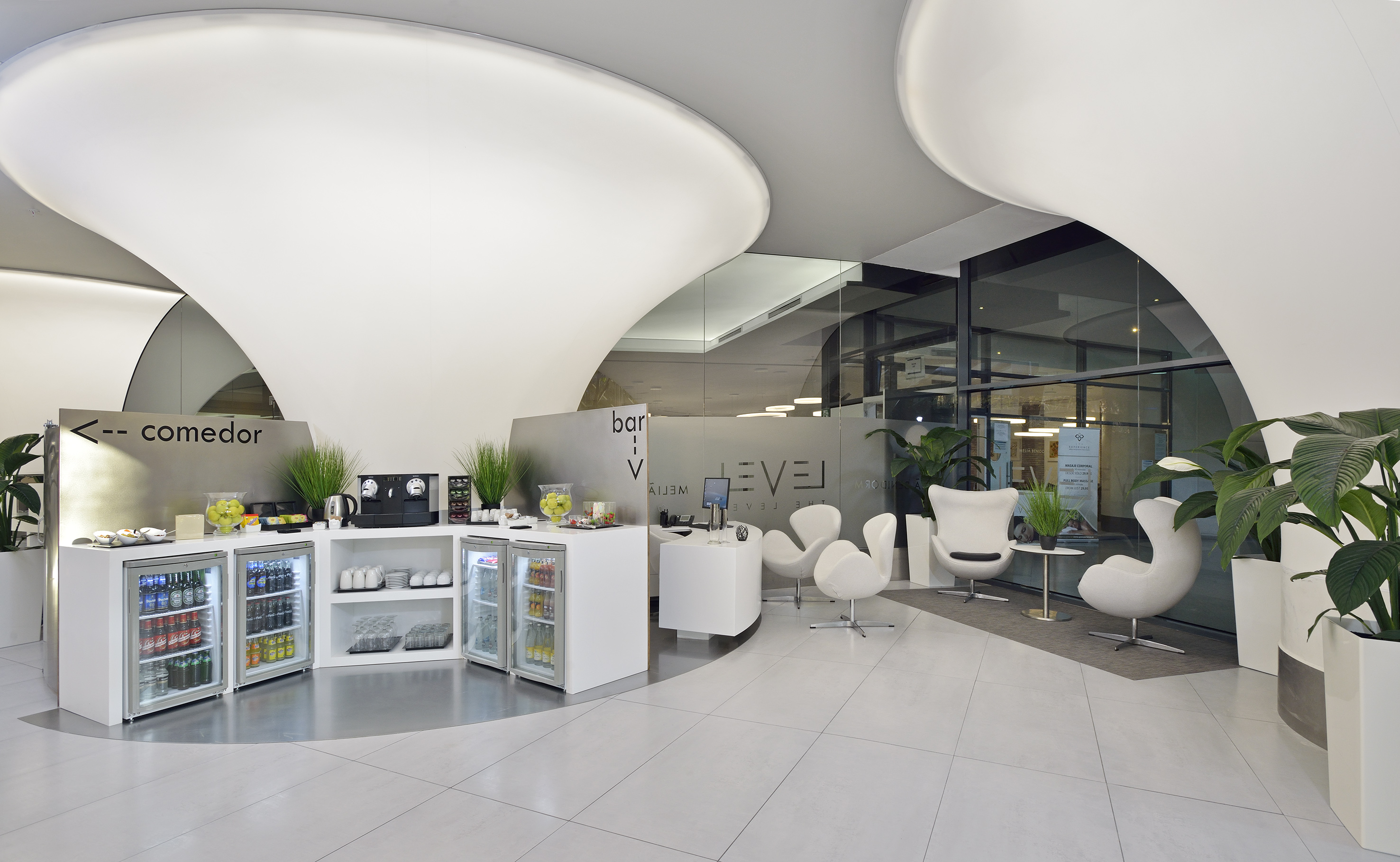 Modern hotel lounge with refreshment station, white armchairs, and plants.