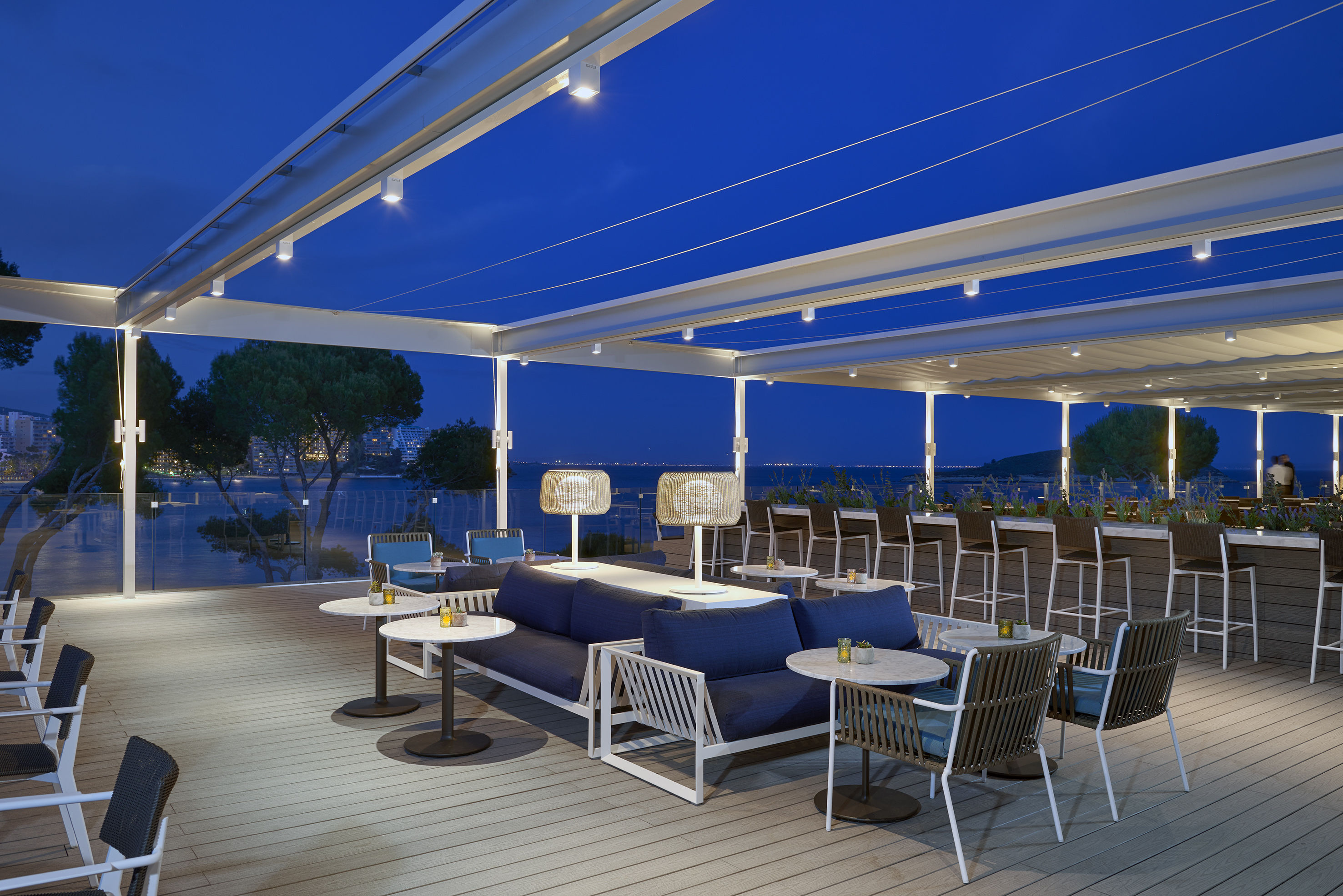Evening outdoor restaurant terrace with modern seating, bar, and lit pergola.