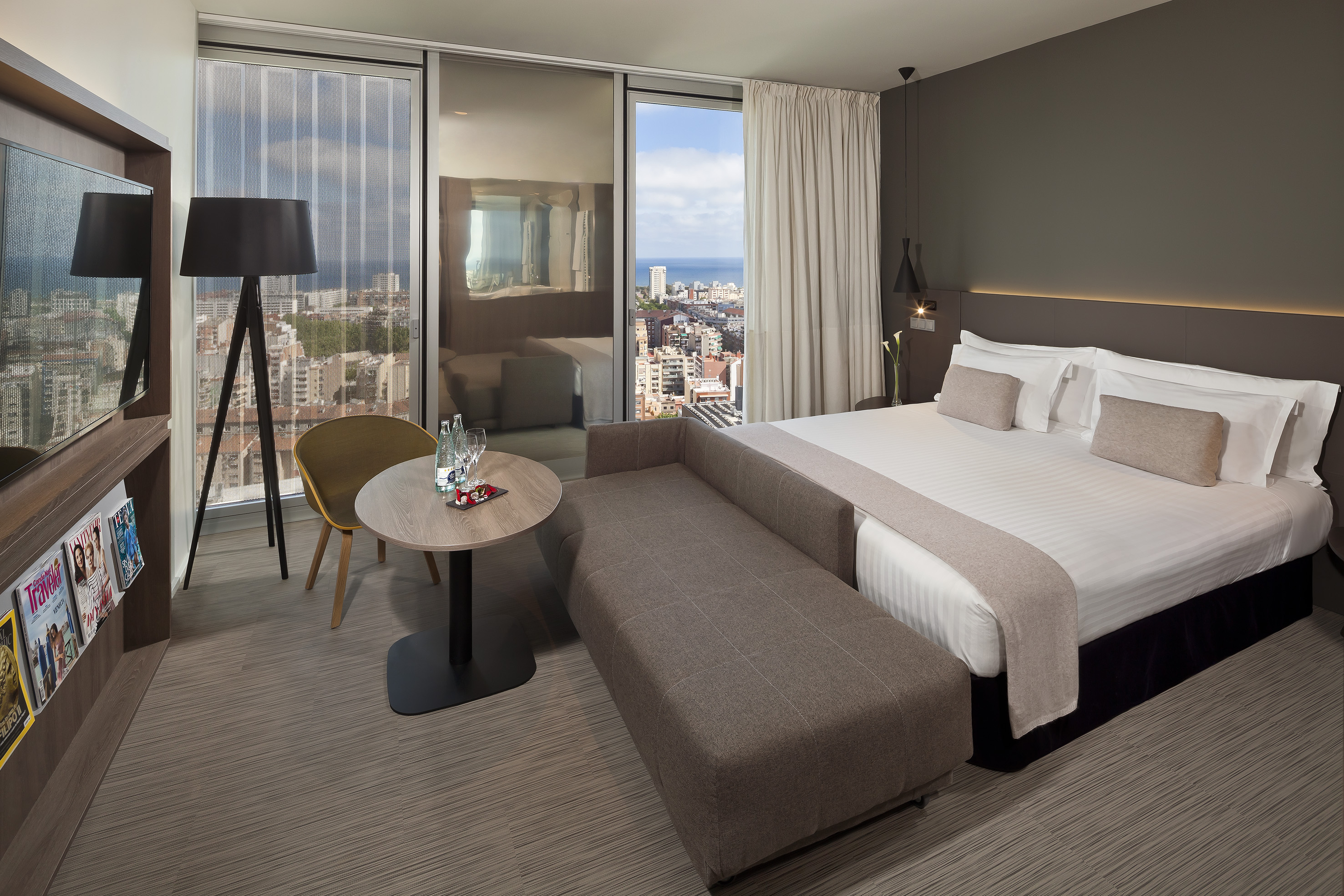 Modern hotel room with large bed, city and ocean view, and contemporary furnishings.