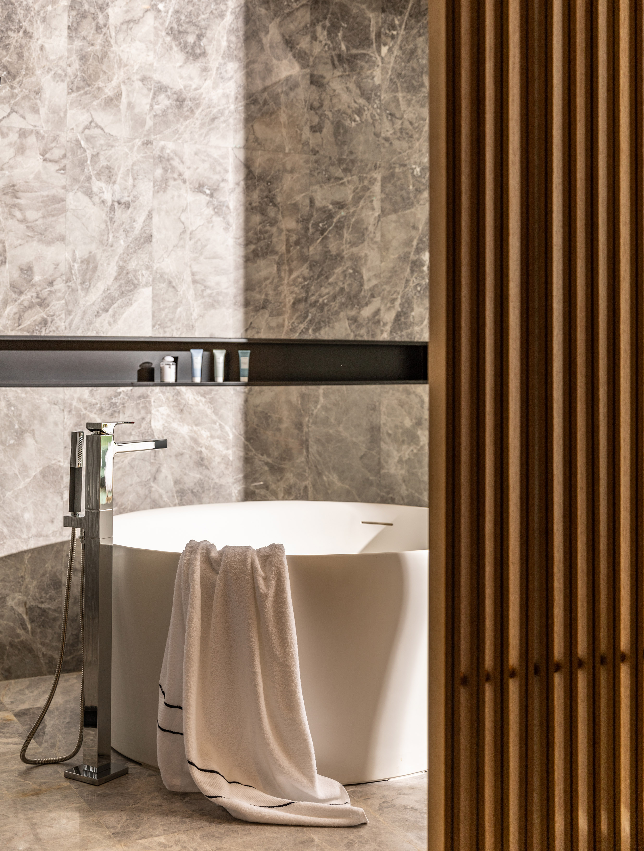 Modern freestanding tub in marble bathroom with chrome faucet and wood screen.