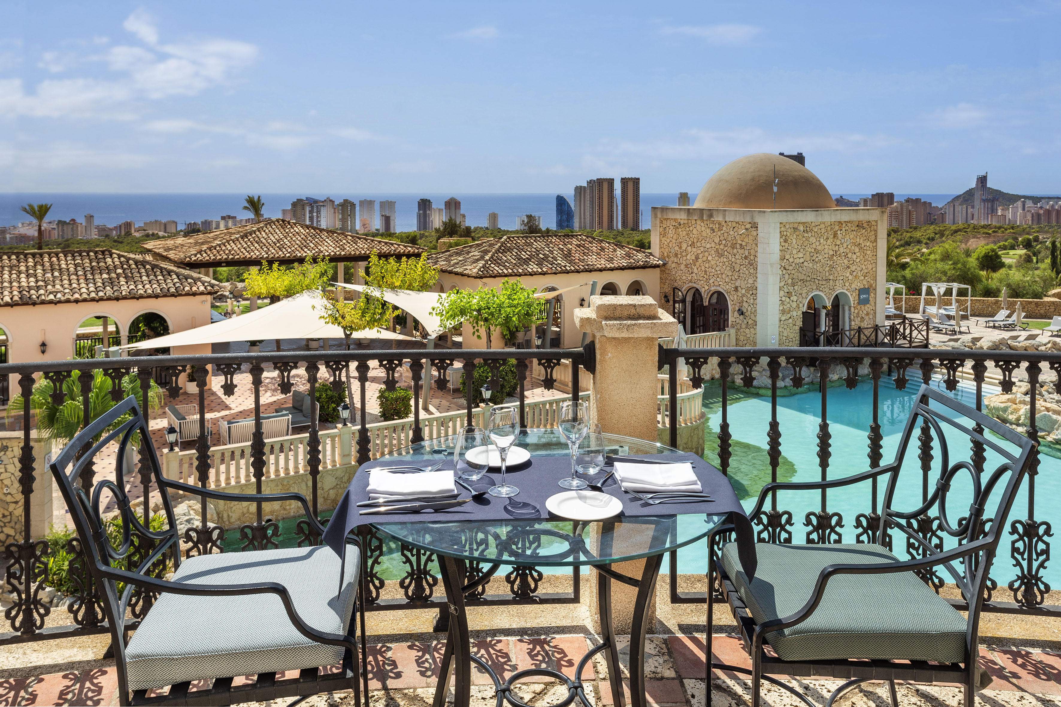 Balcony dining table overlooking a resort, pool, sea, and city skyline.