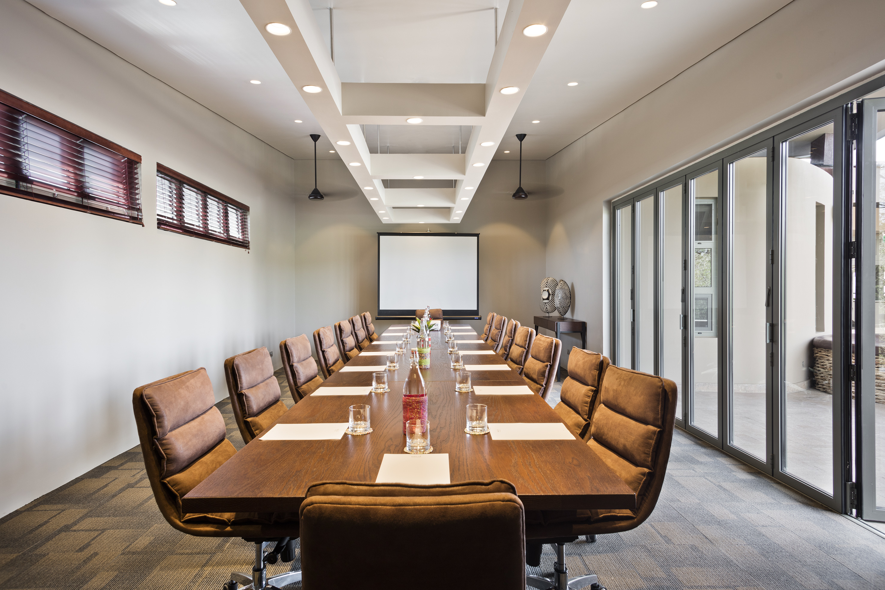 Spacious boardroom with long table, brown chairs, projector screen, and glass doors.