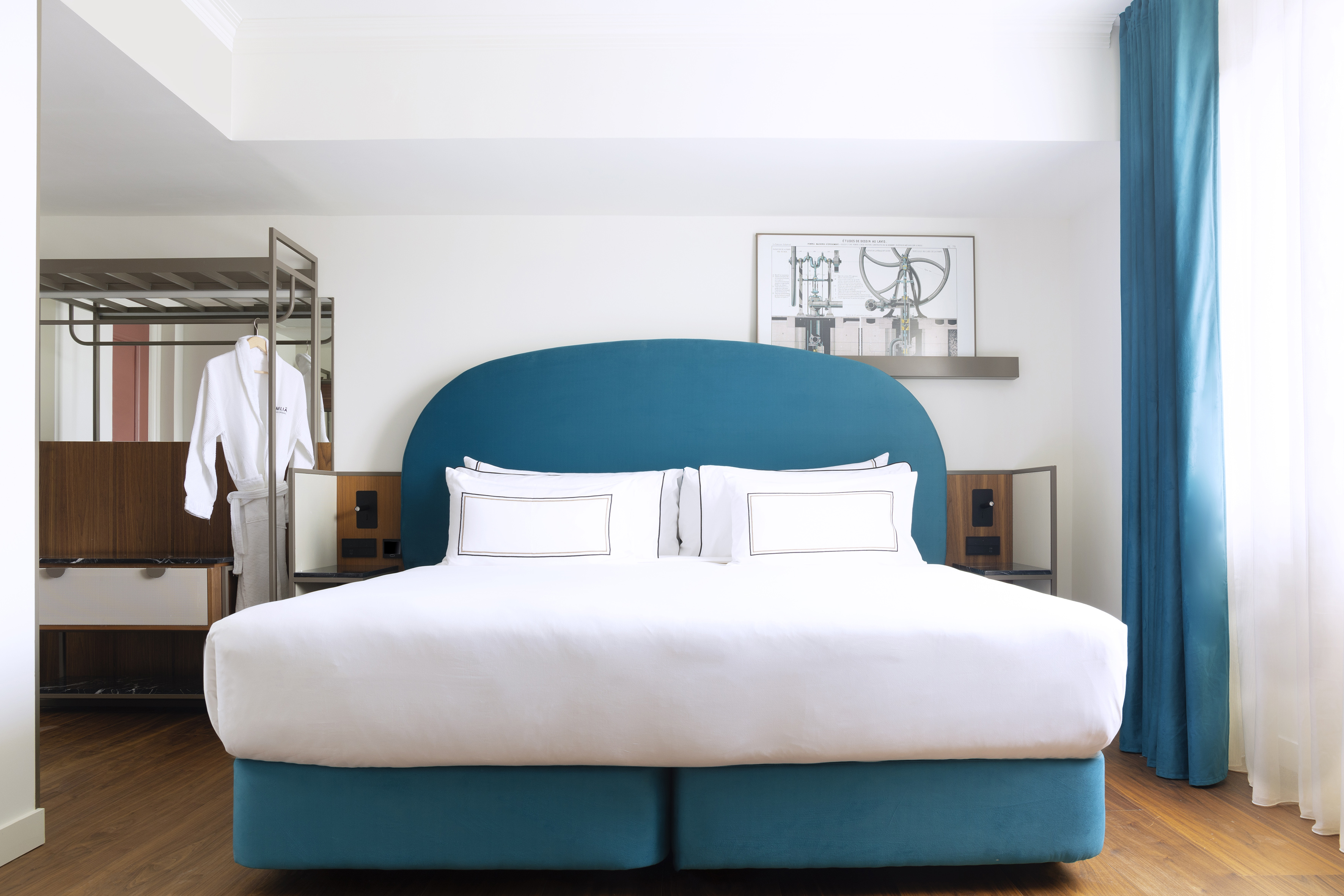 Modern hotel room with teal king bed, white linens, wood floor, and bathrobe.
