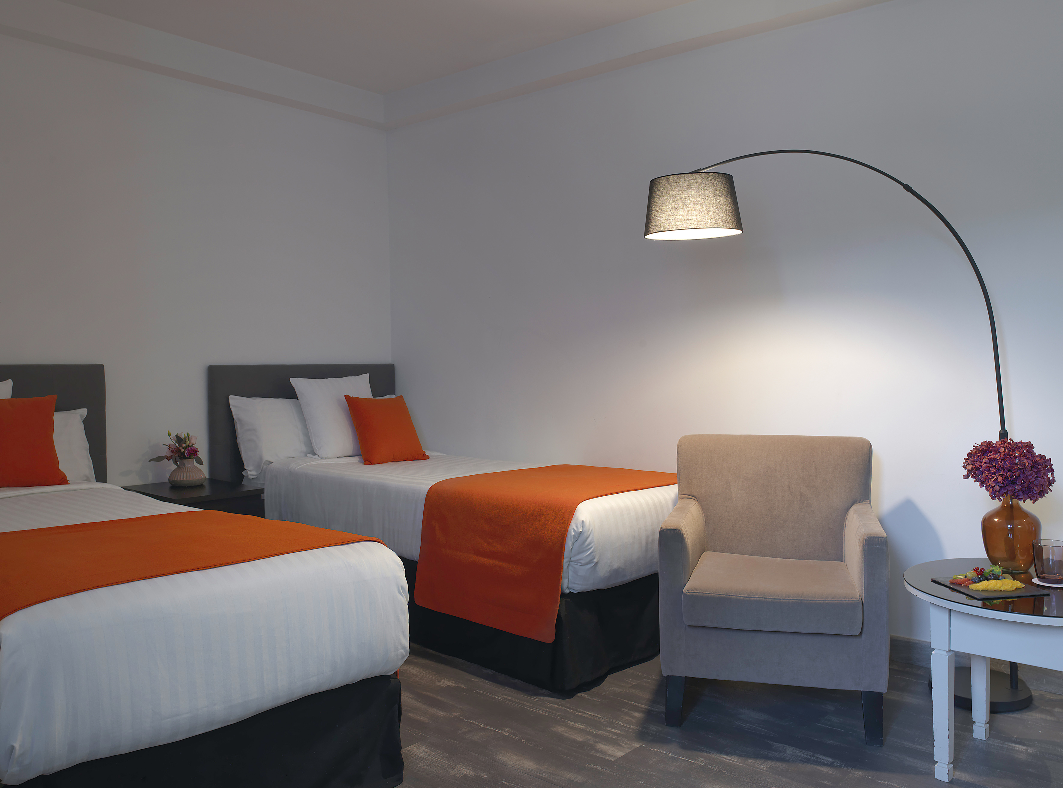 Twin hotel room with orange accents, grey armchair, and modern arc lamp.