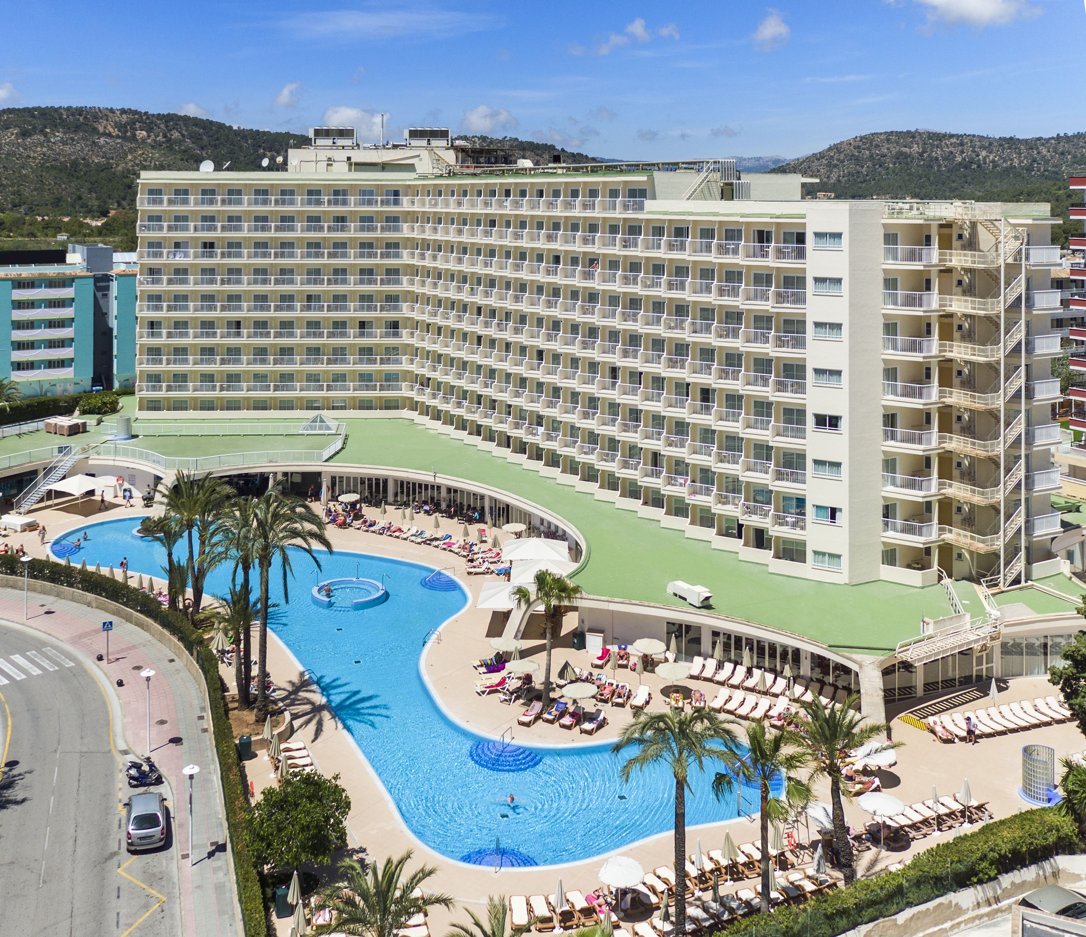 Sunny resort hotel with large swimming pool, palm trees, and lounge area.