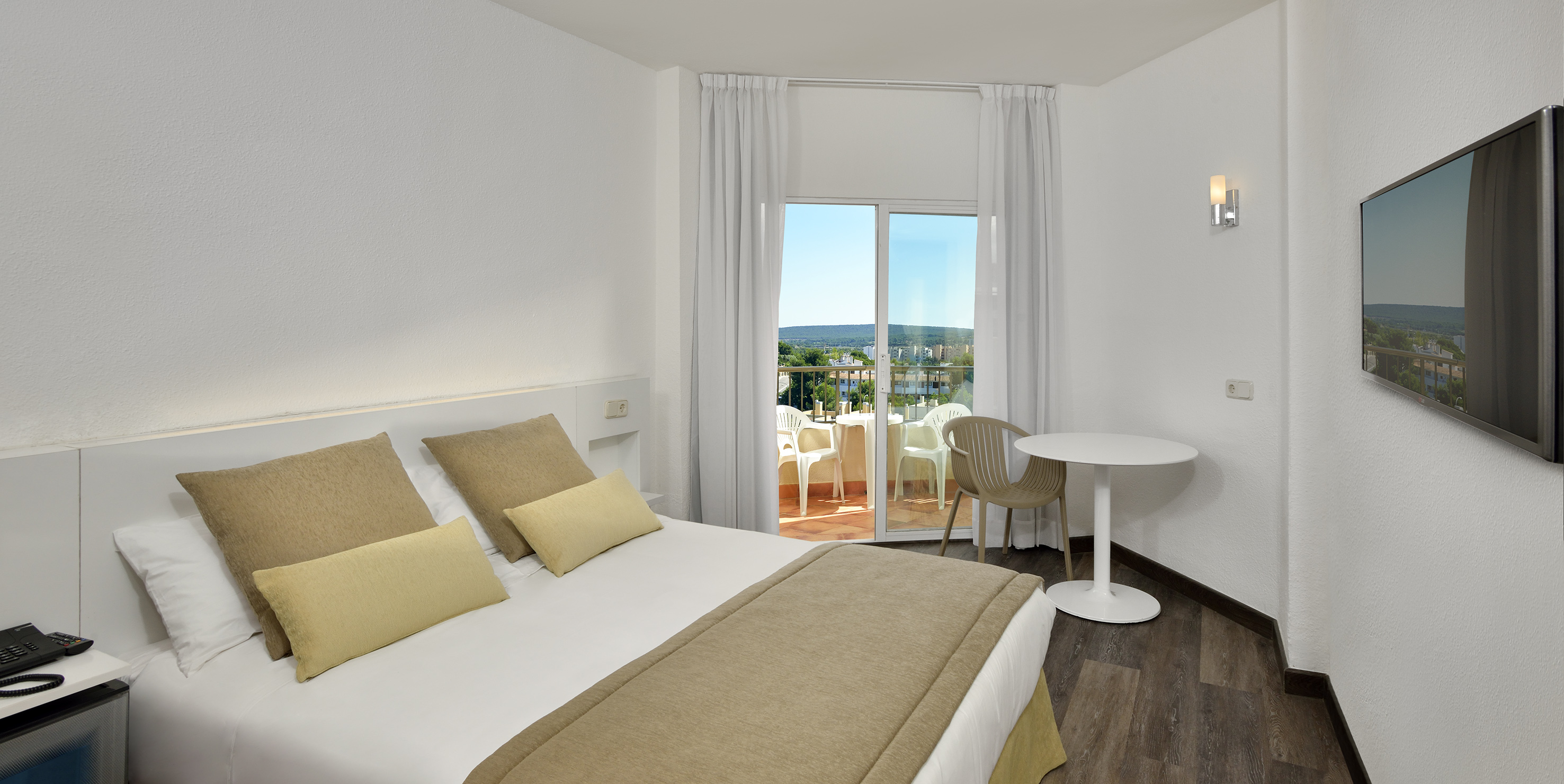 Bright hotel room with bed, balcony, outdoor seating, and scenic view.