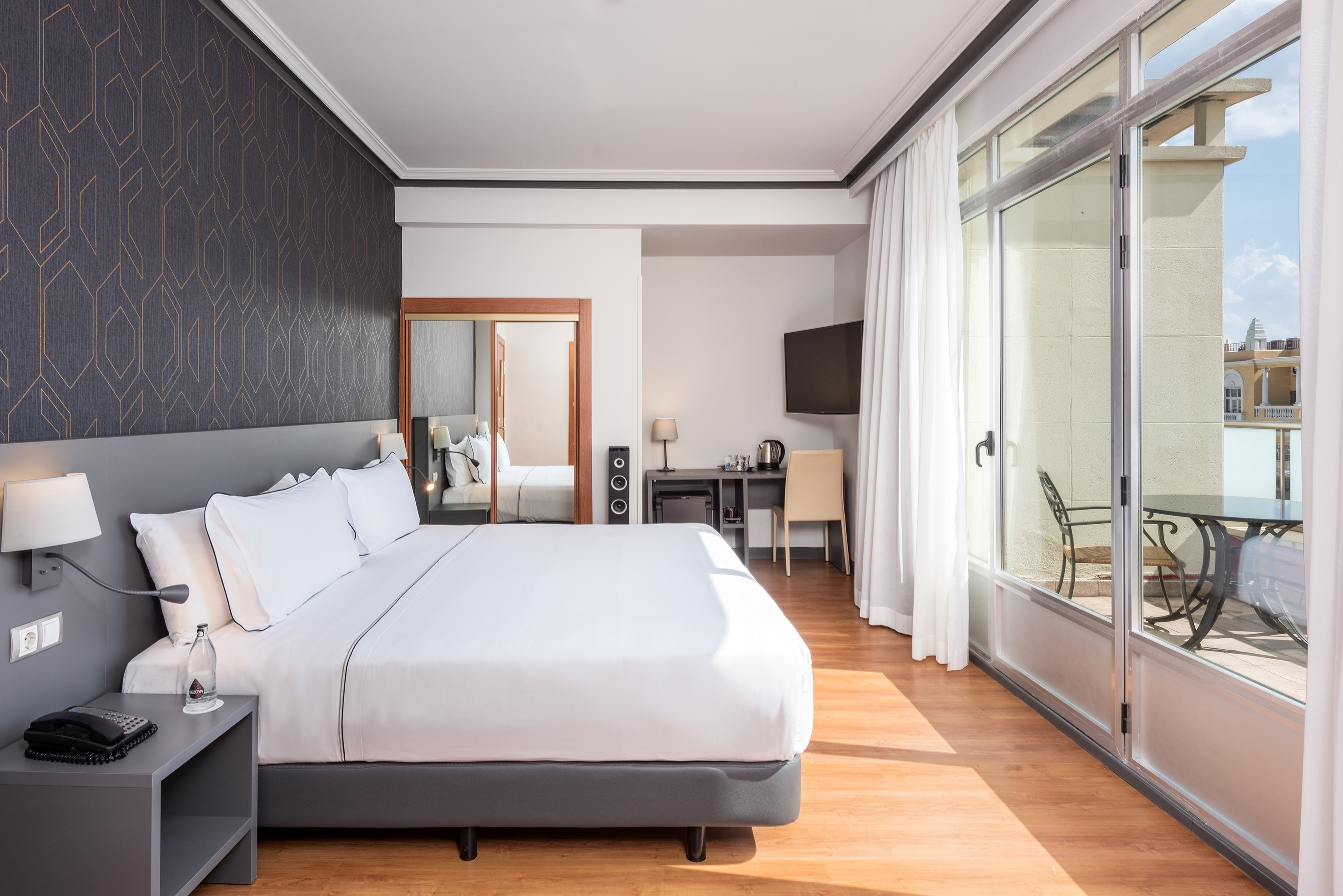 Modern hotel room with king bed, geometric accent wall, desk, and balcony.