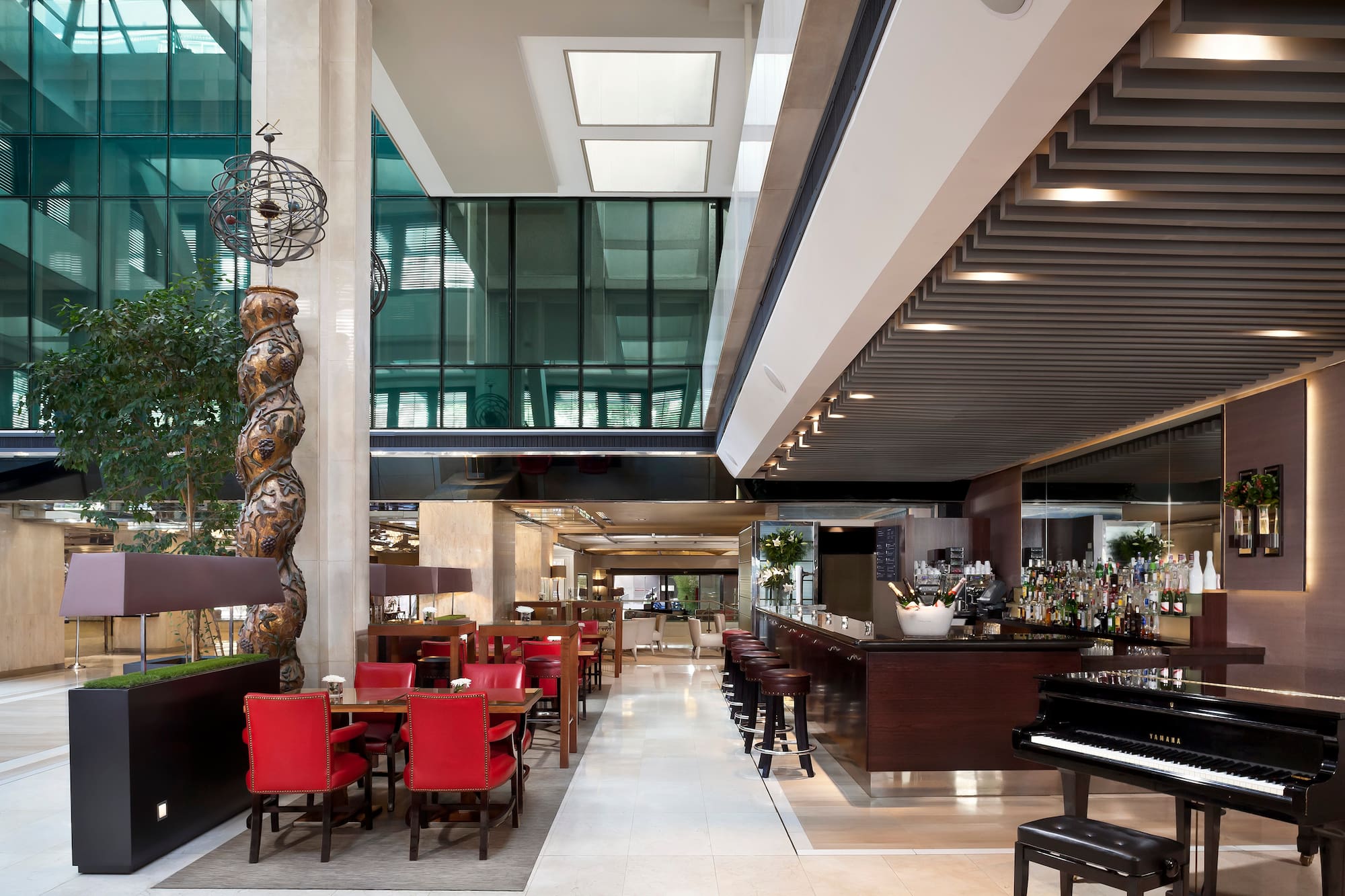 Modern hotel bar and lounge, red chairs, grand piano, glass windows.
