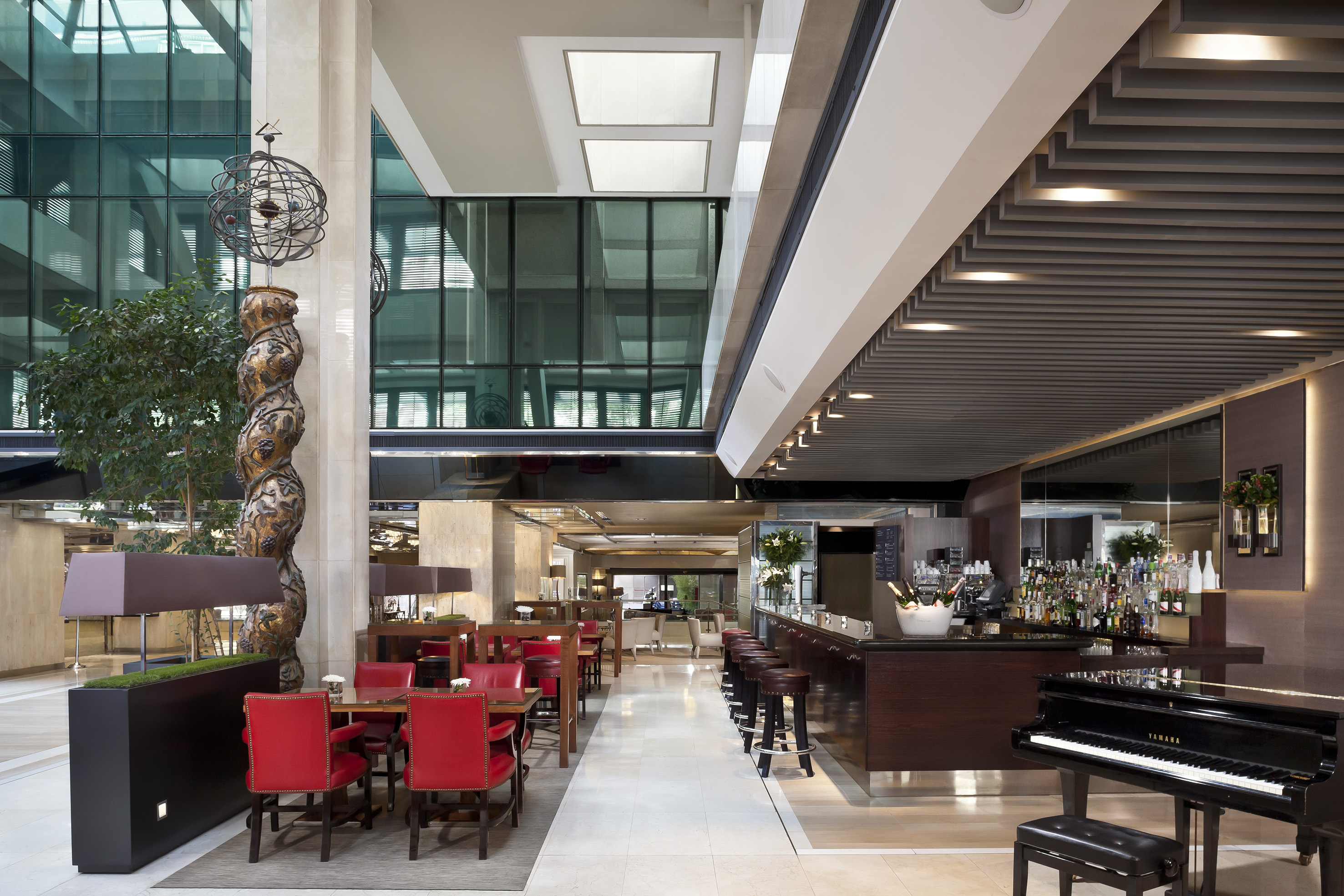 Modern hotel bar and lounge, red chairs, grand piano, glass windows.