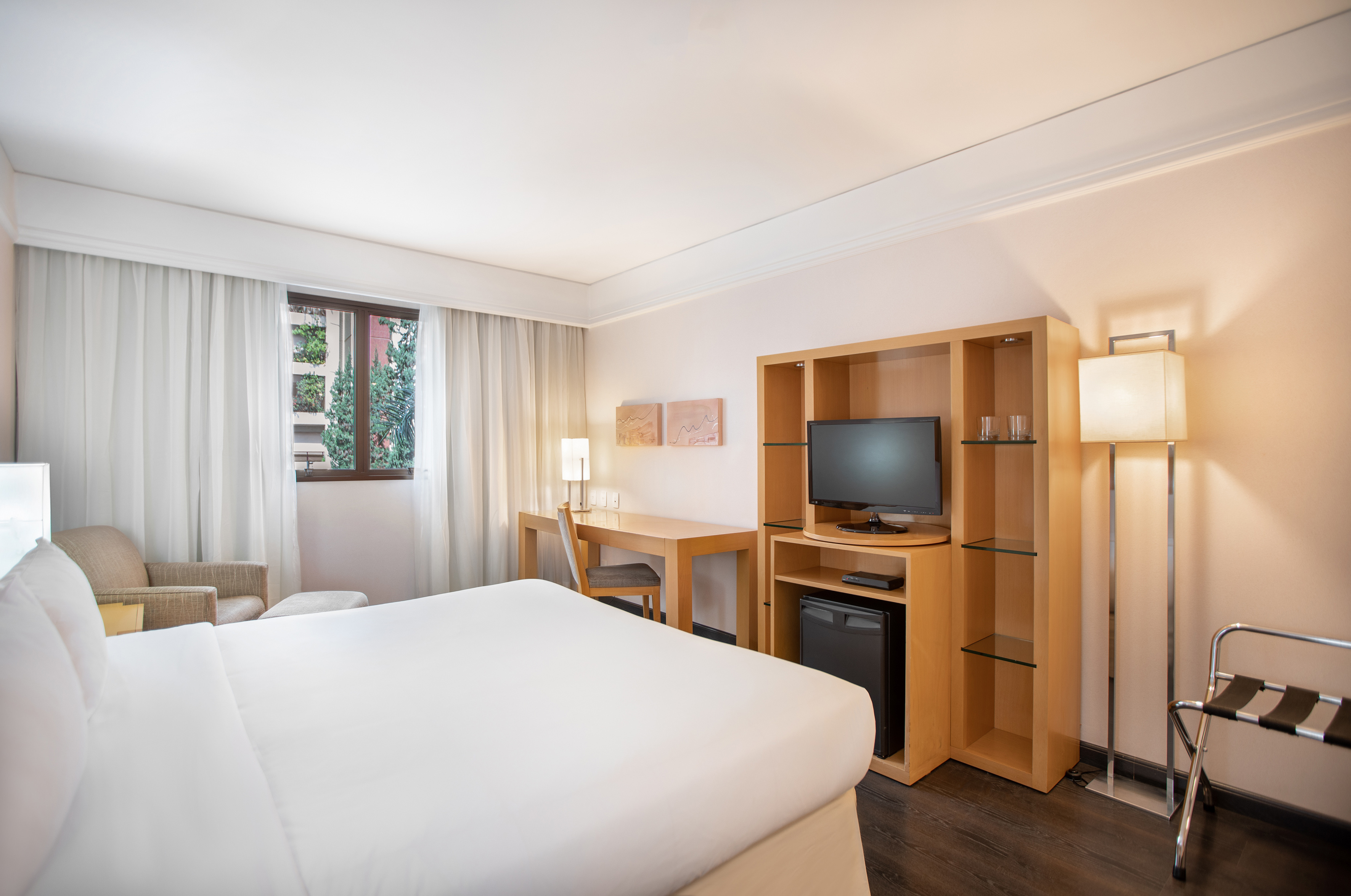 Modern hotel guest room with bed, desk, TV, window, and mini-fridge.