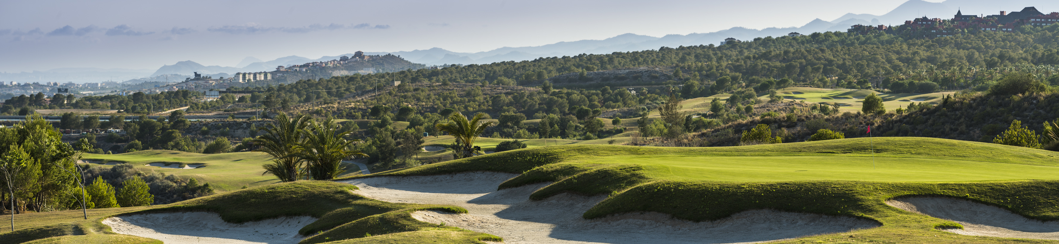 Green golf course with sand bunkers, palm trees, and distant mountains.