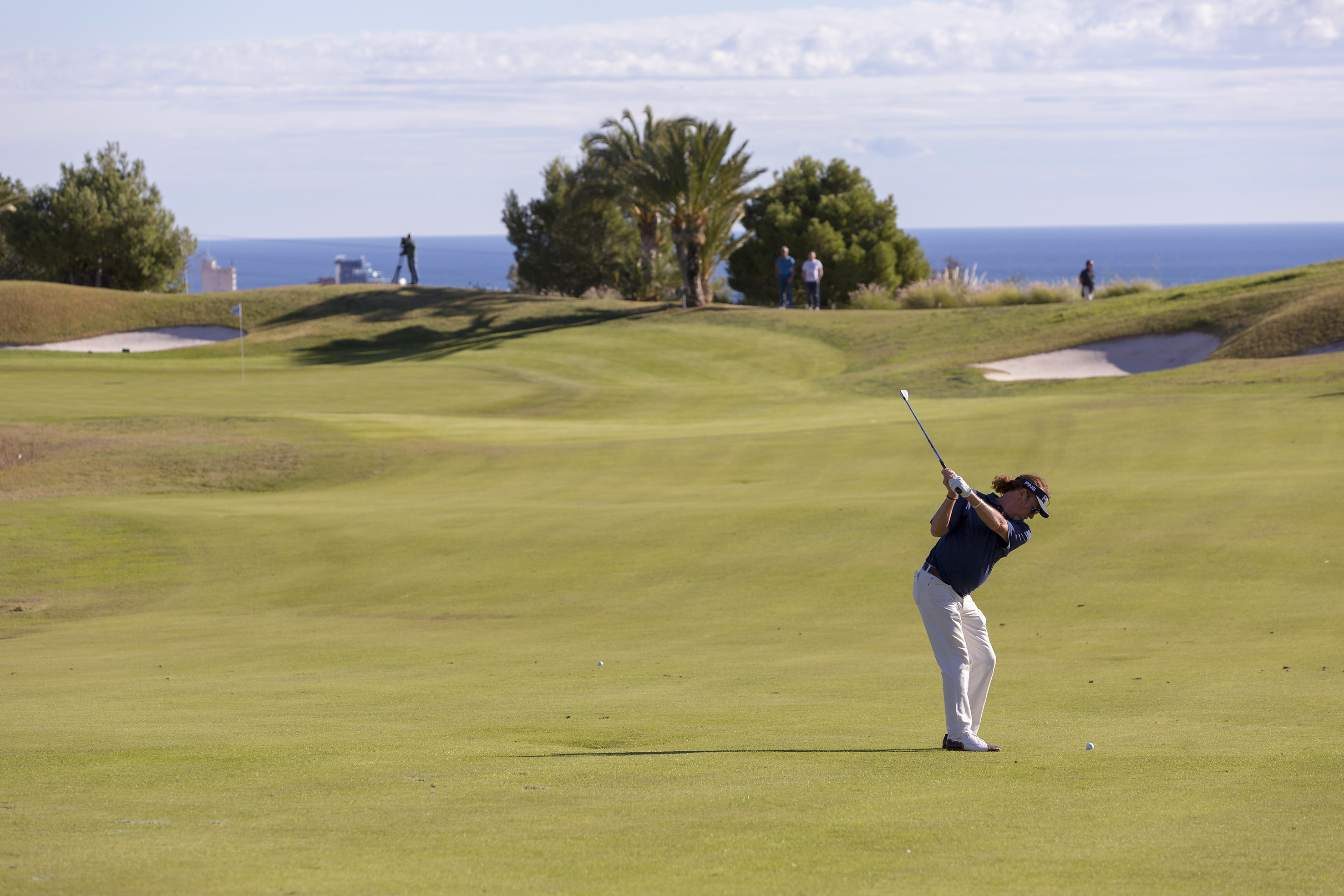 Golfer mid-swing on a green coastal golf course with ocean views.