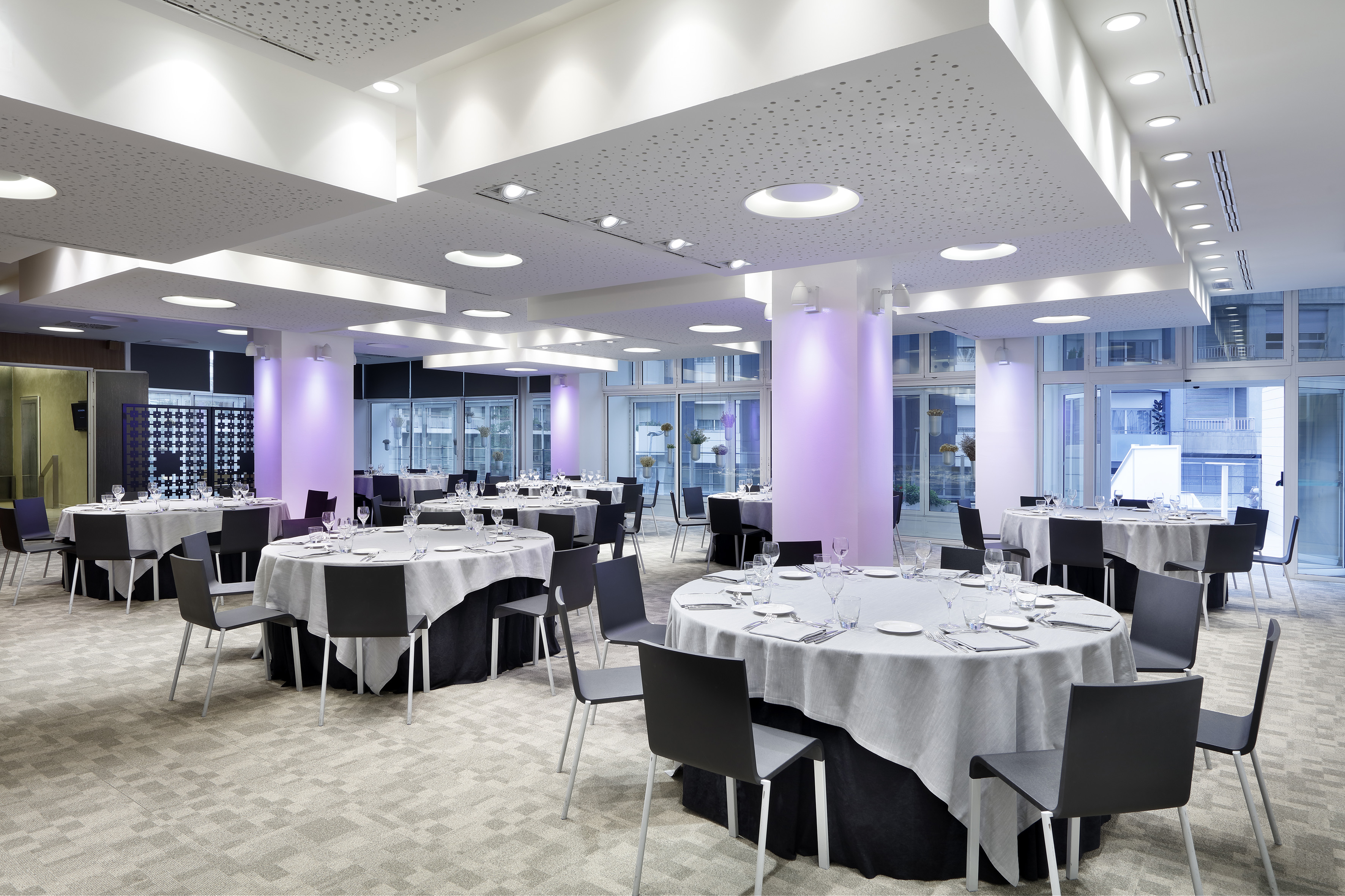 Modern banquet hall with round tables, dark chairs, and purple lighting.