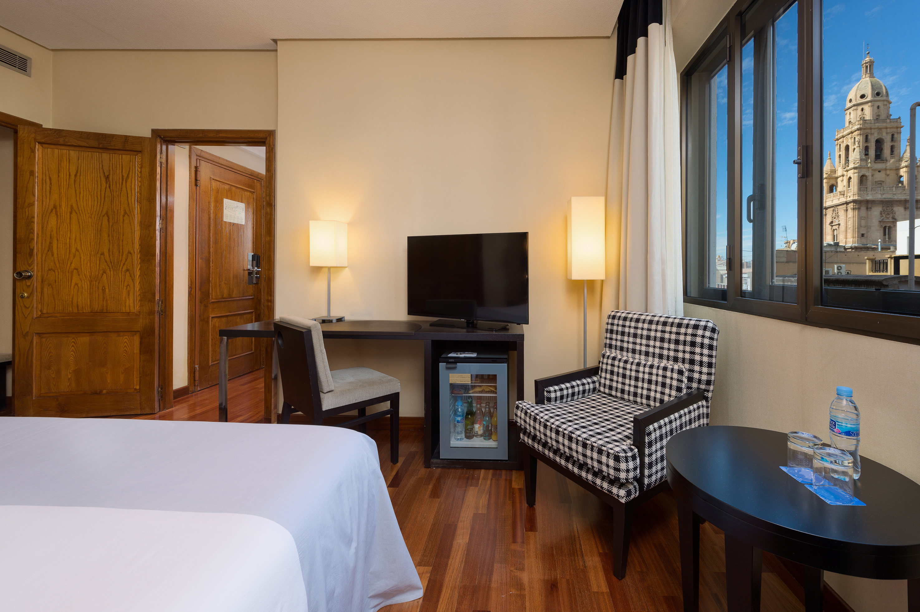 Hotel room with bed, desk, TV, armchair, and window view of Murcia Cathedral.