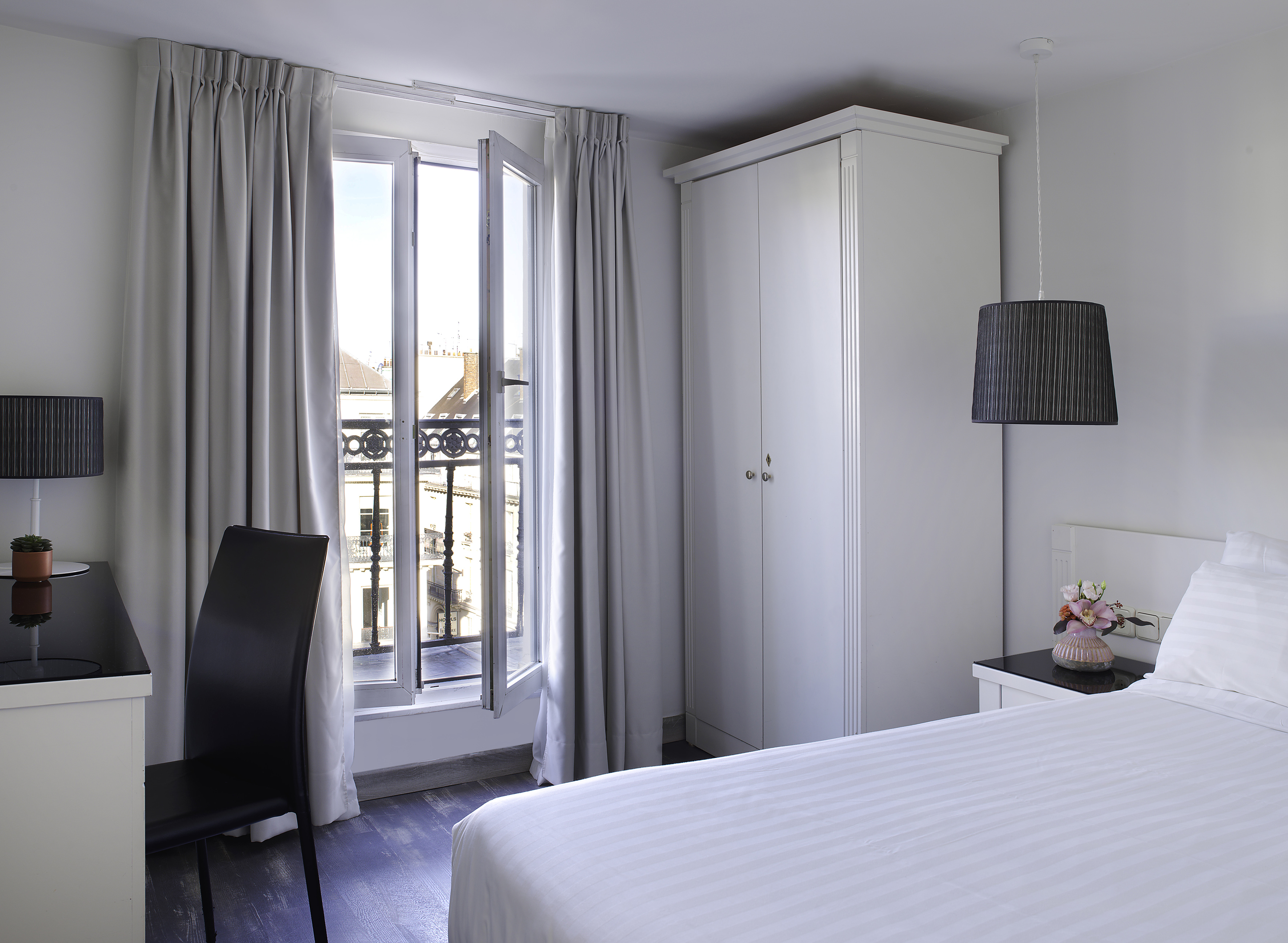 Bright hotel room with white bed, open French window, grey curtains, and wardrobe.