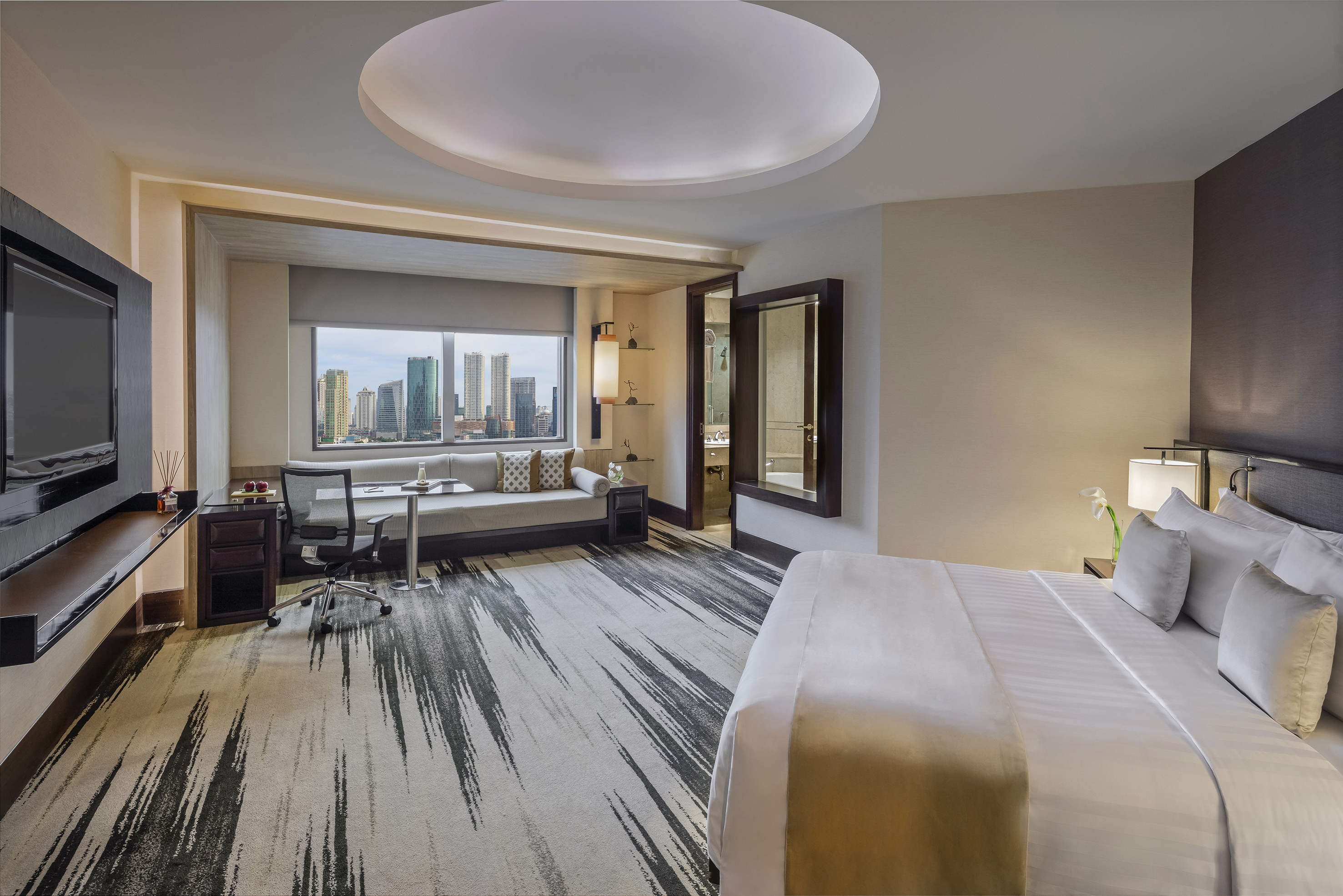 Luxury hotel suite with city view, king bed, desk, and abstract carpet.