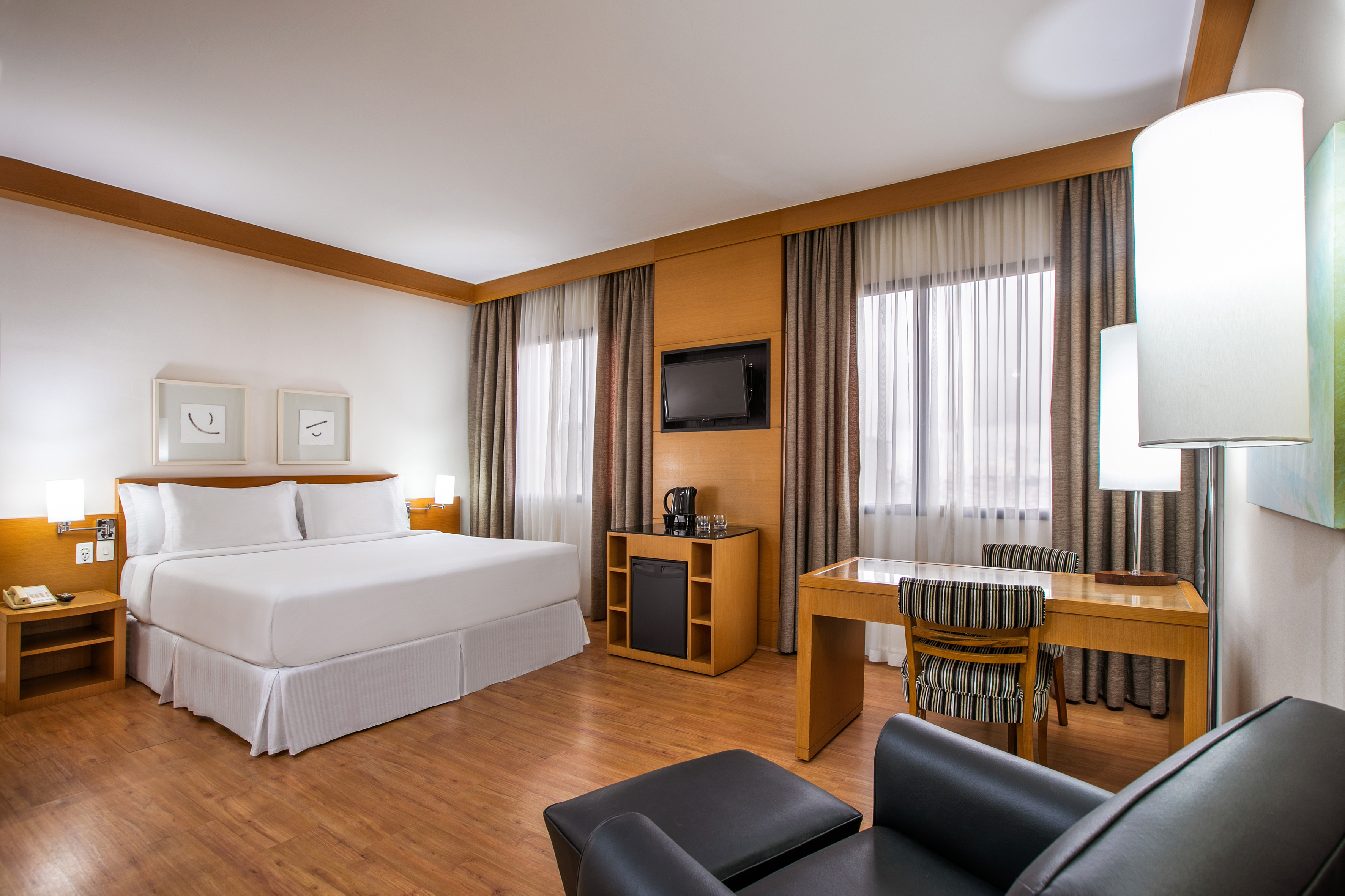 Modern hotel room with large bed, wooden desk, TV, and warm wood flooring.