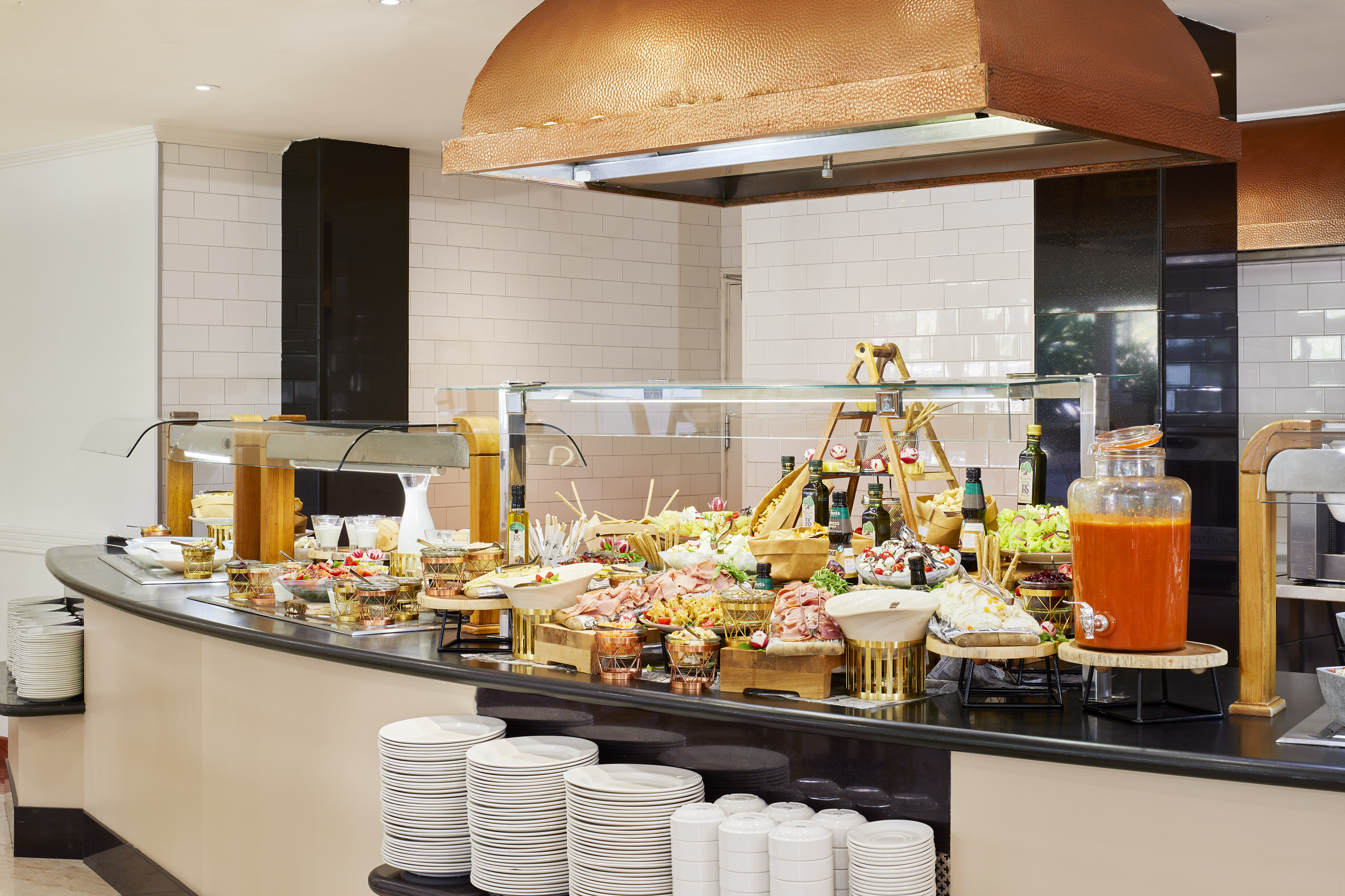 Restaurant buffet with cold cuts, fresh salads, juice, and stacked plates.