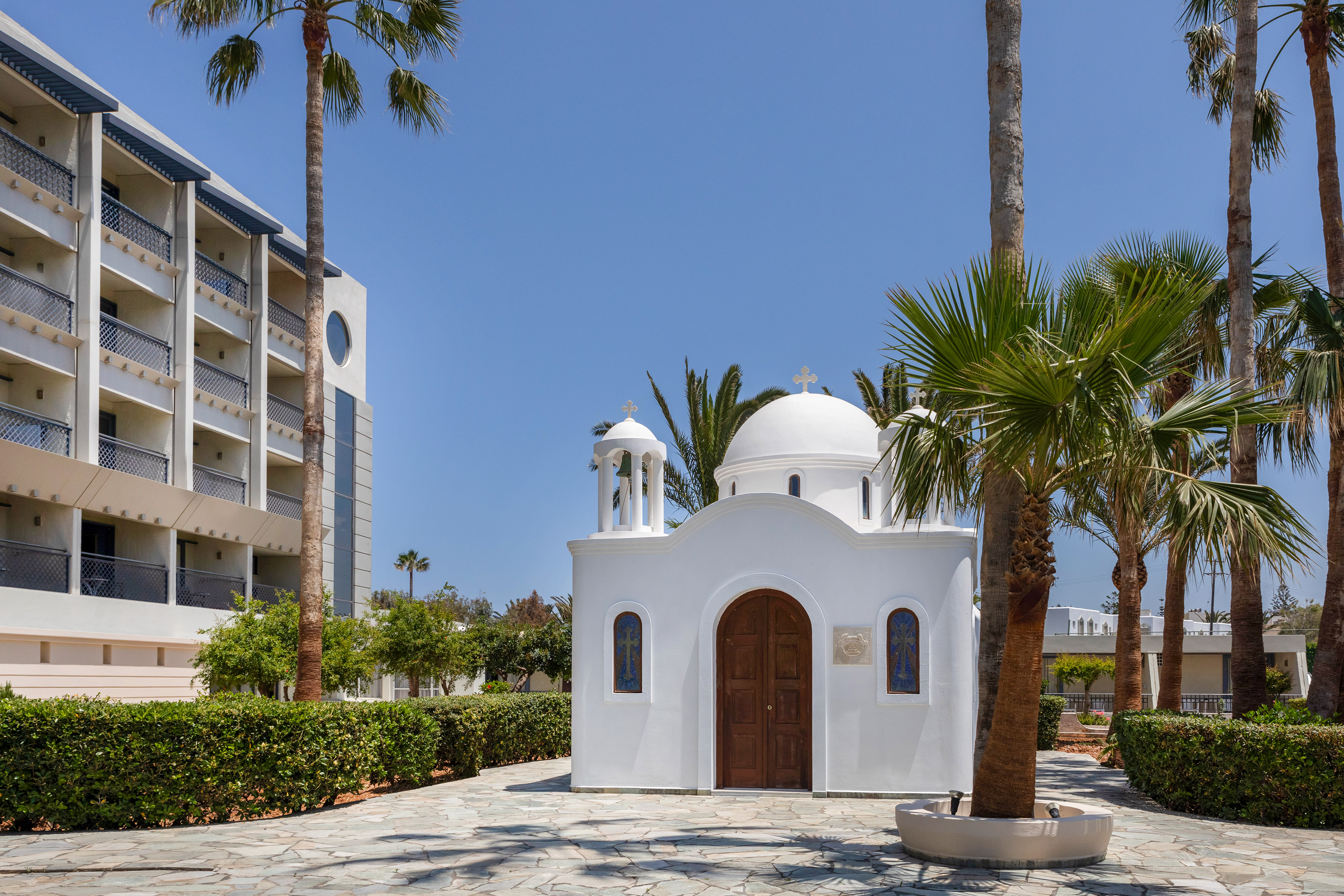 White domed chapel, bell tower, palm trees, and modern hotel building.