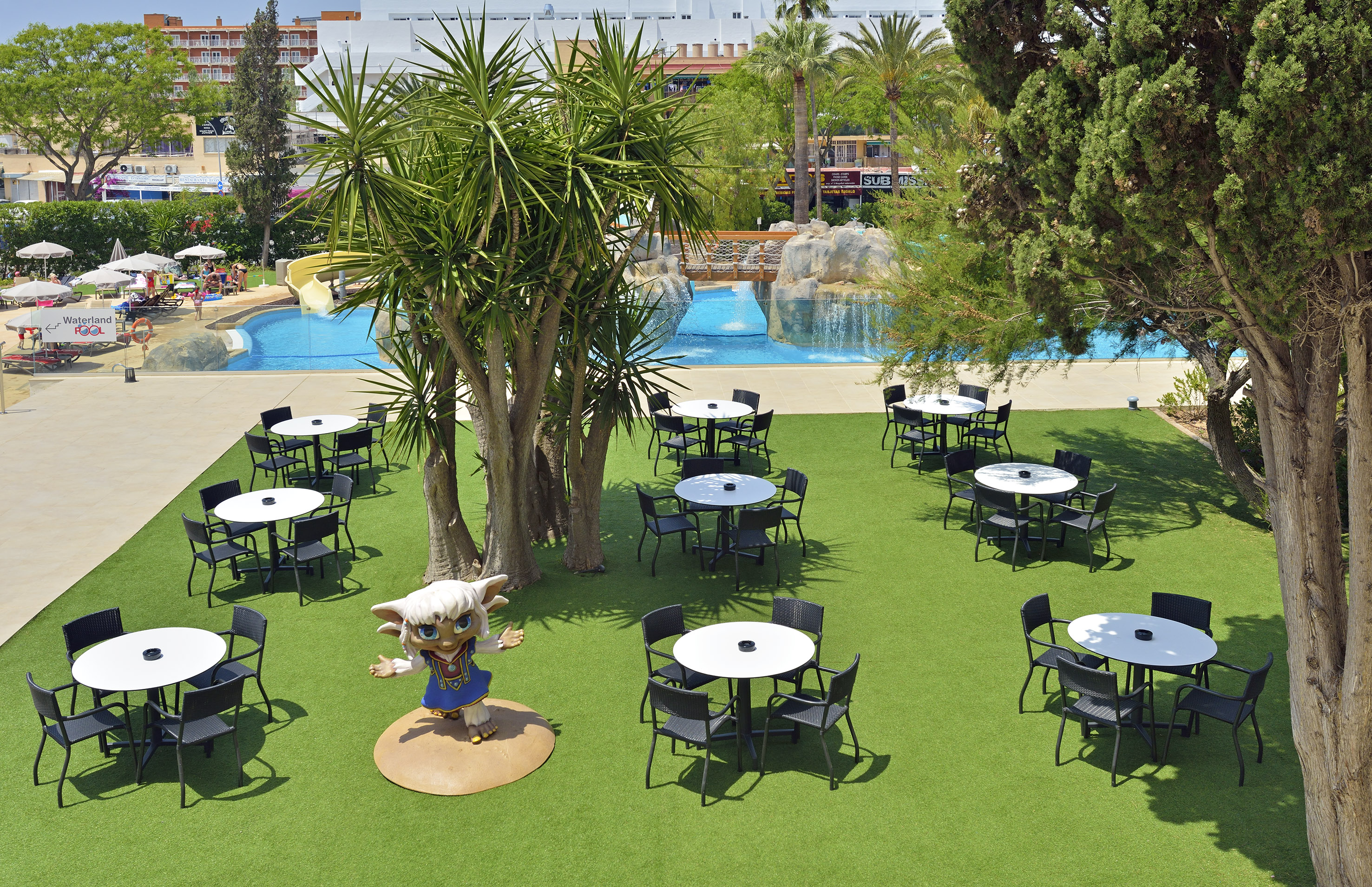 Sunny resort pool with tables on artificial grass, water slide, and mascot.