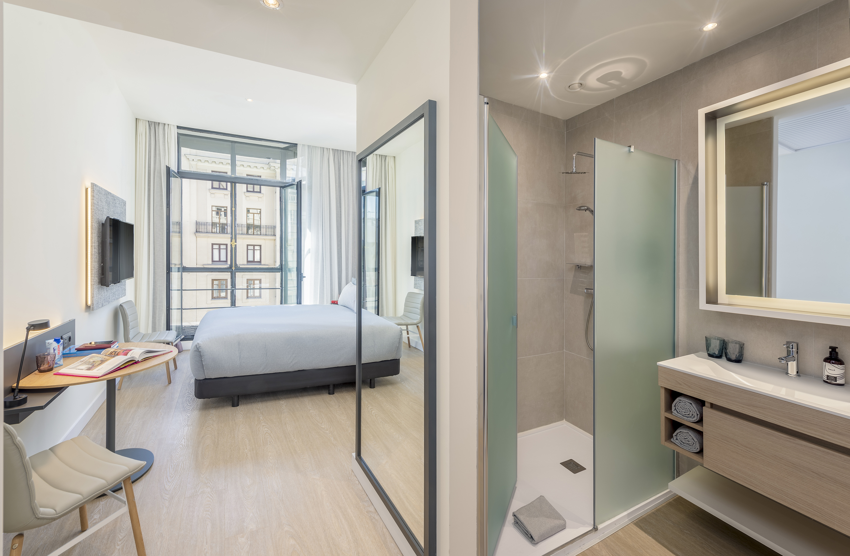 Modern hotel room with bed, city view window, and integrated bathroom shower.