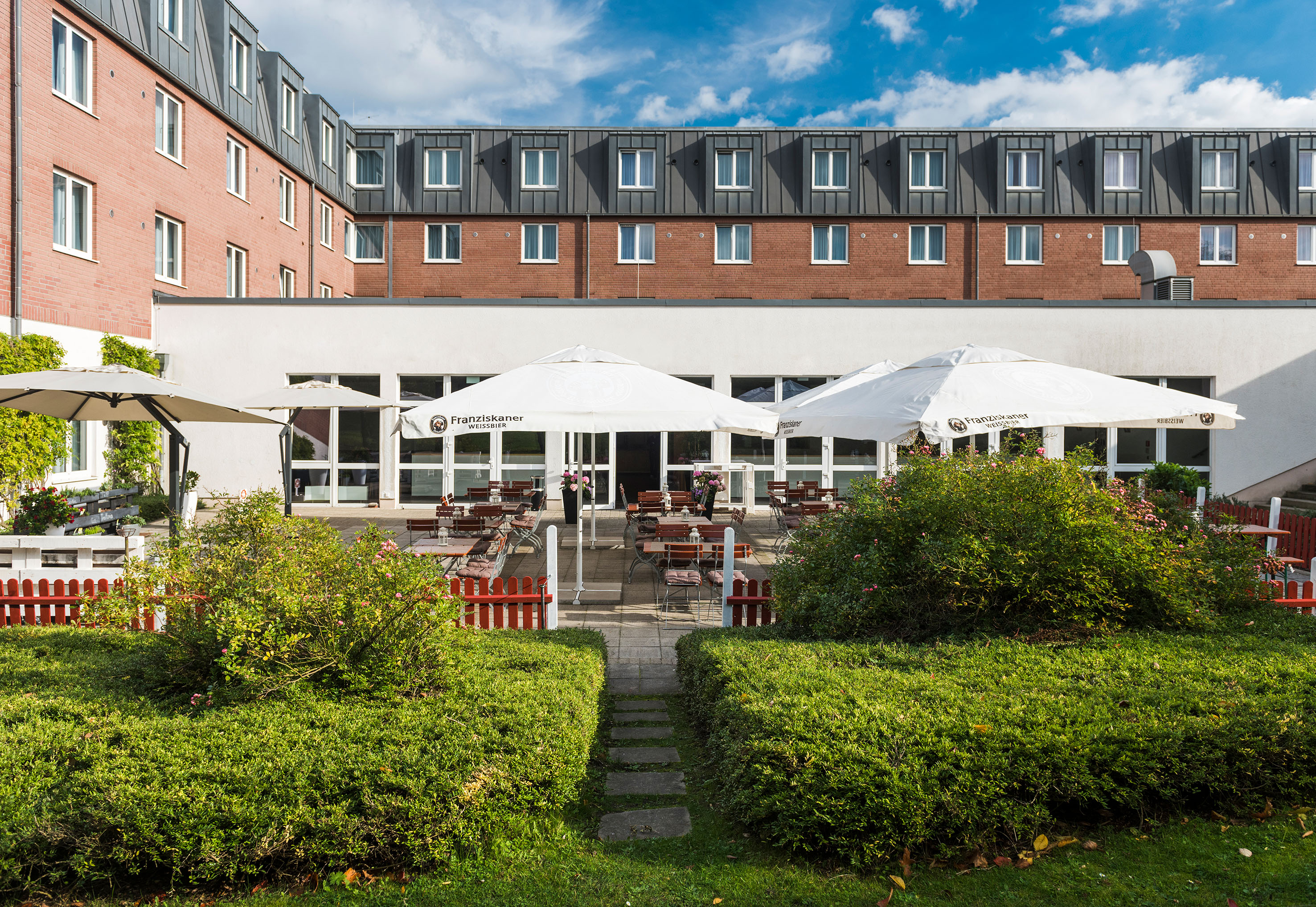 Hotel building with outdoor patio, Franziskaner umbrellas, and green garden.