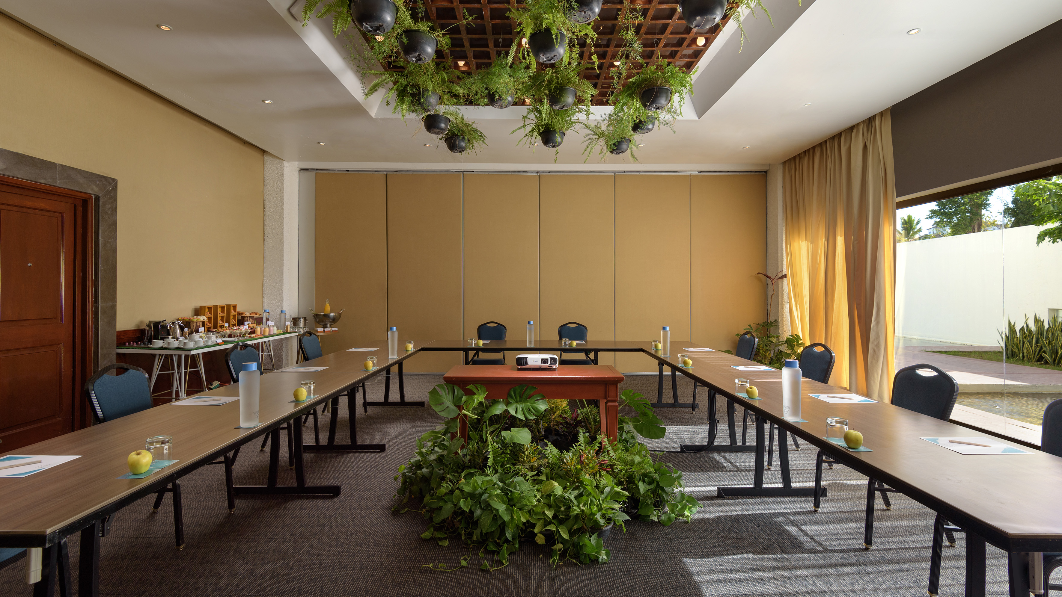 Conference room with U-shaped setup, projector, refreshments, and lush plants.