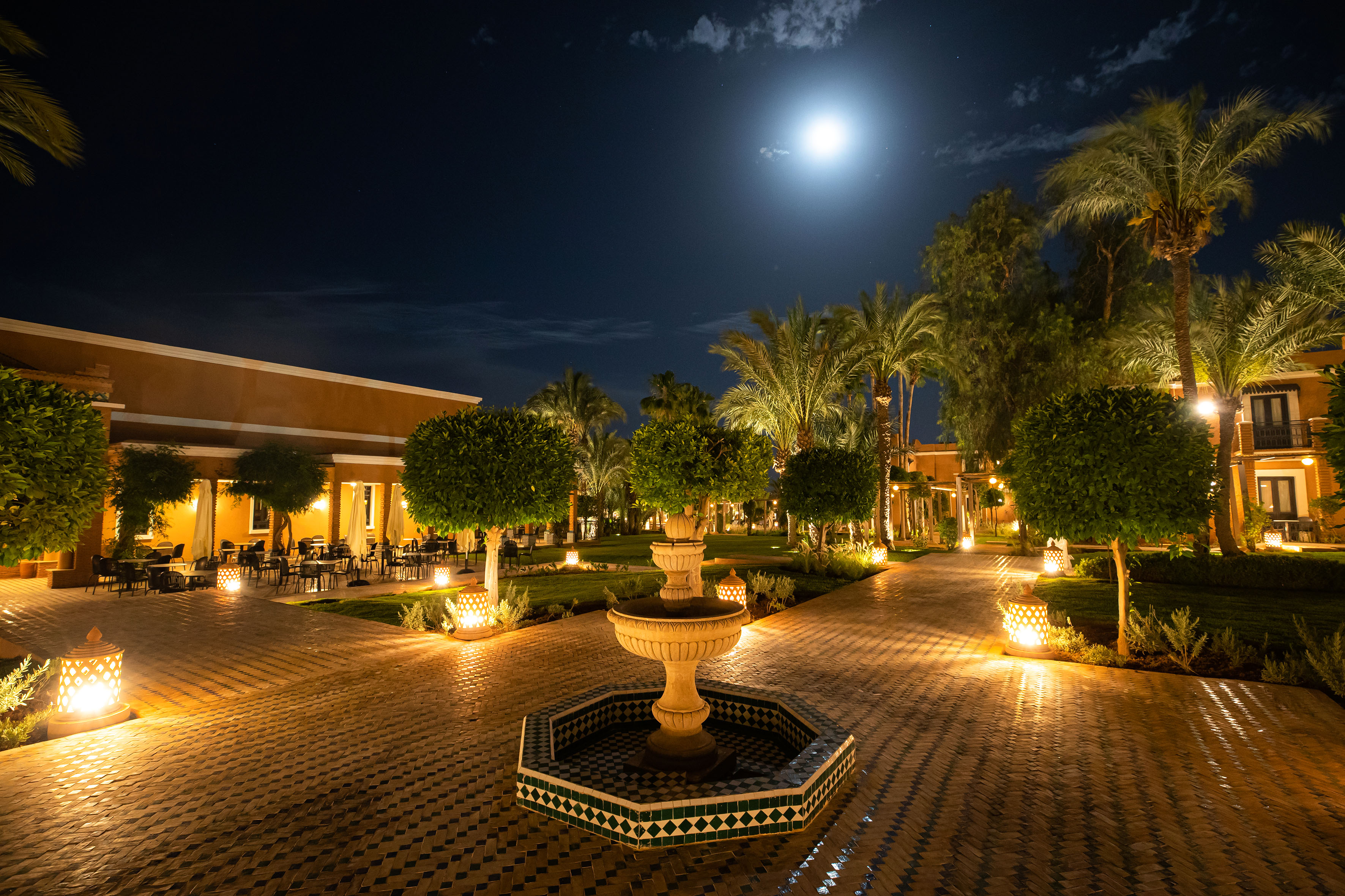 Illuminated Moroccan resort garden at night with moon, palm trees, and fountain.