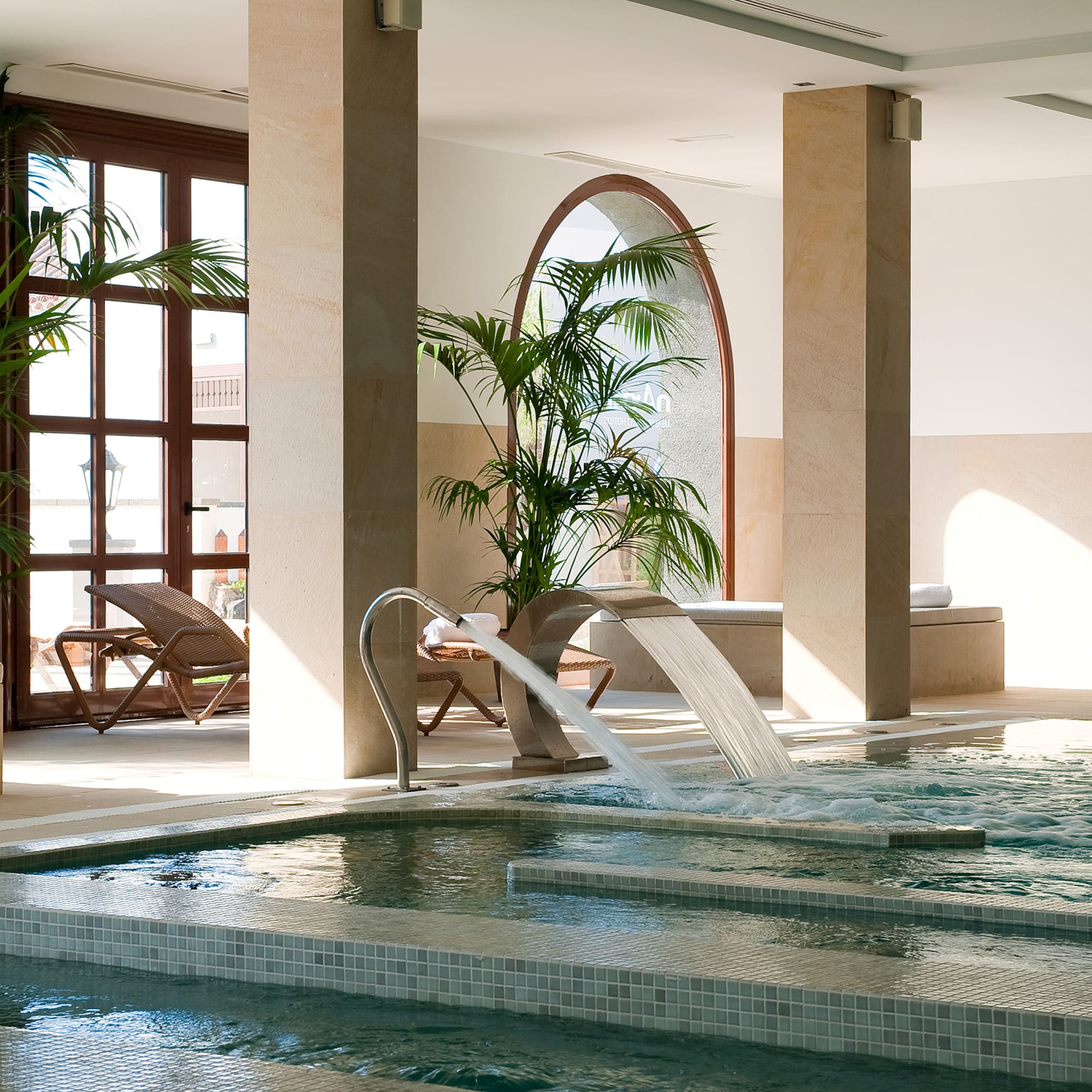 Luxury spa indoor pool with waterfall, palm plants, and woman in bathrobe.