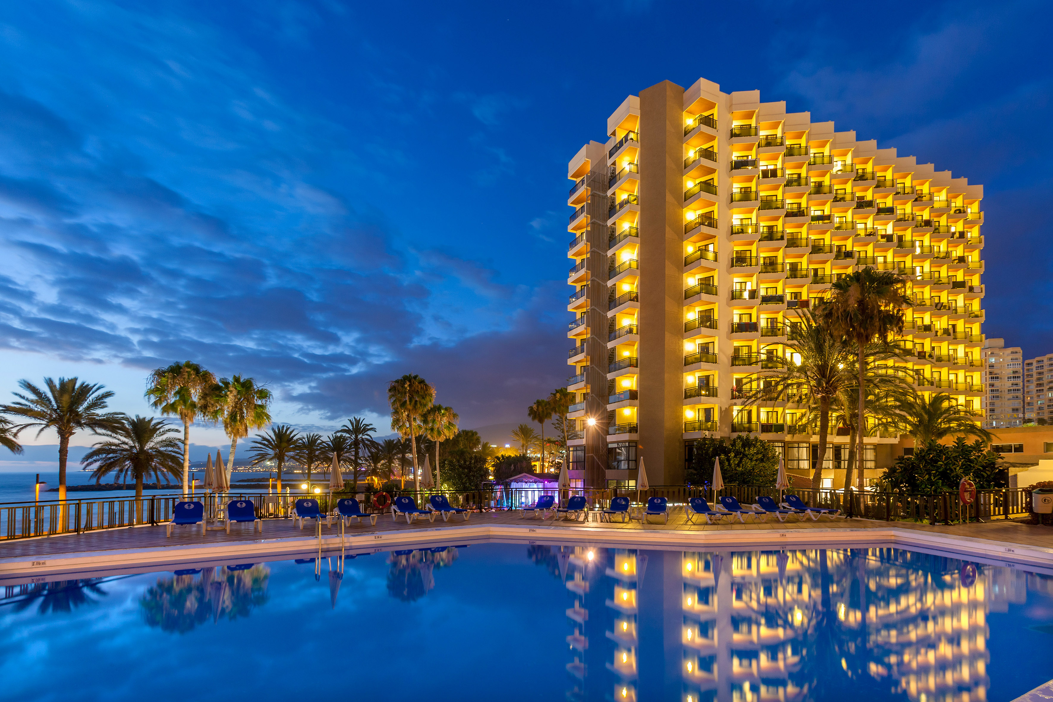 Resort hotel with illuminated pool, palm trees, and ocean at dusk.