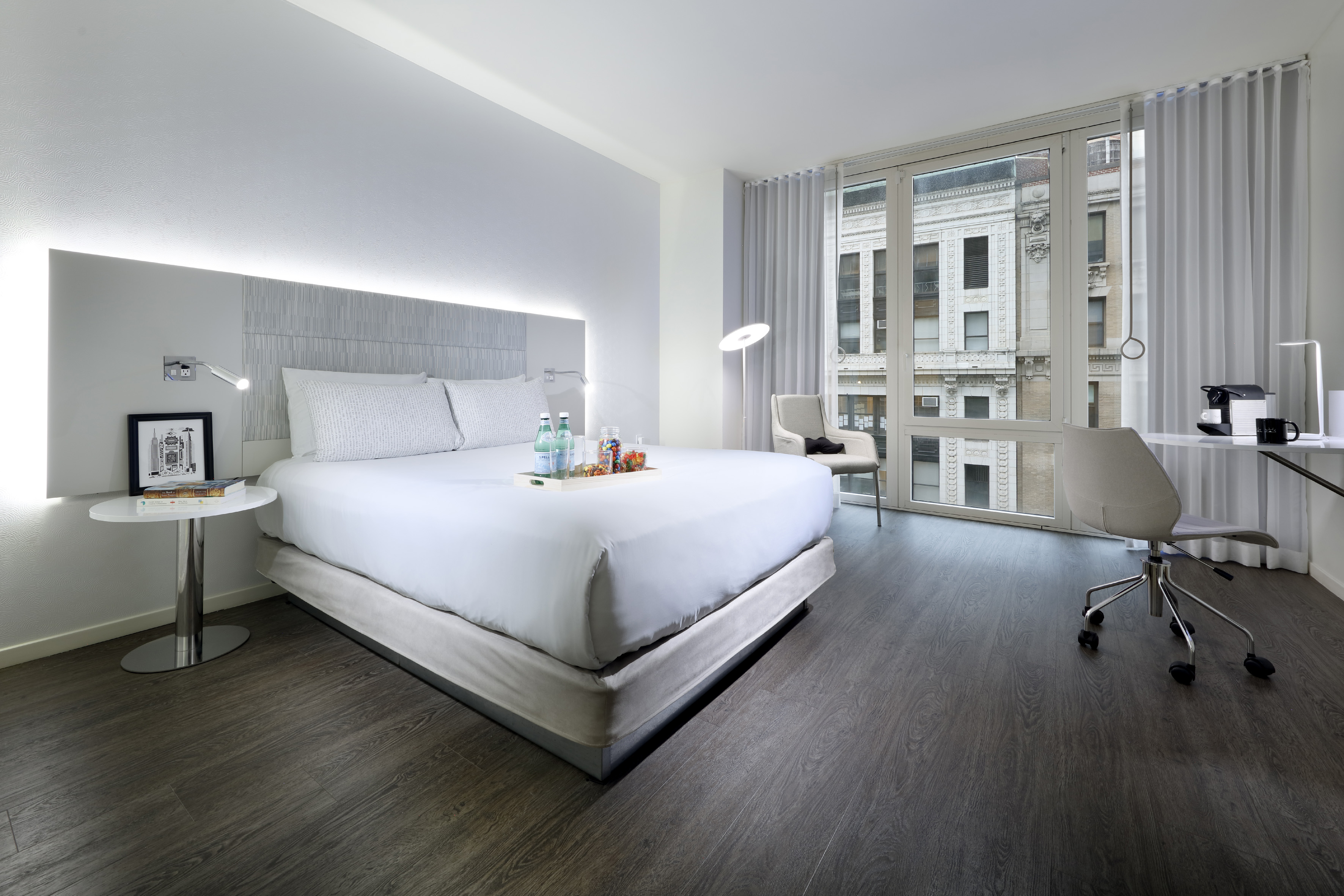 Modern hotel room with king bed, city view, desk, coffee machine, dark floor.