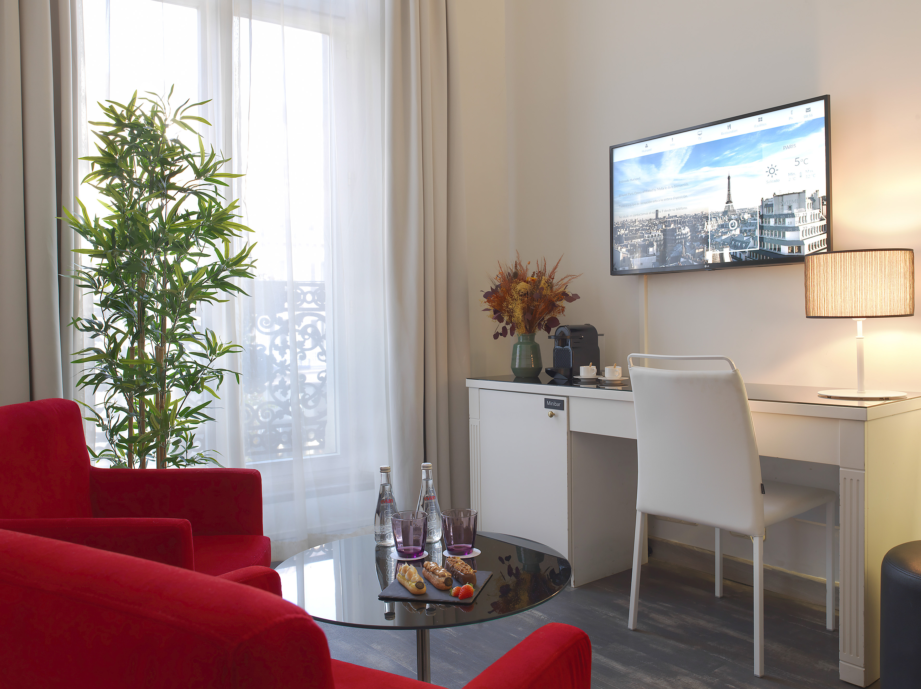 Luxurious hotel room with red armchairs, desk, TV showing Paris, and minibar.