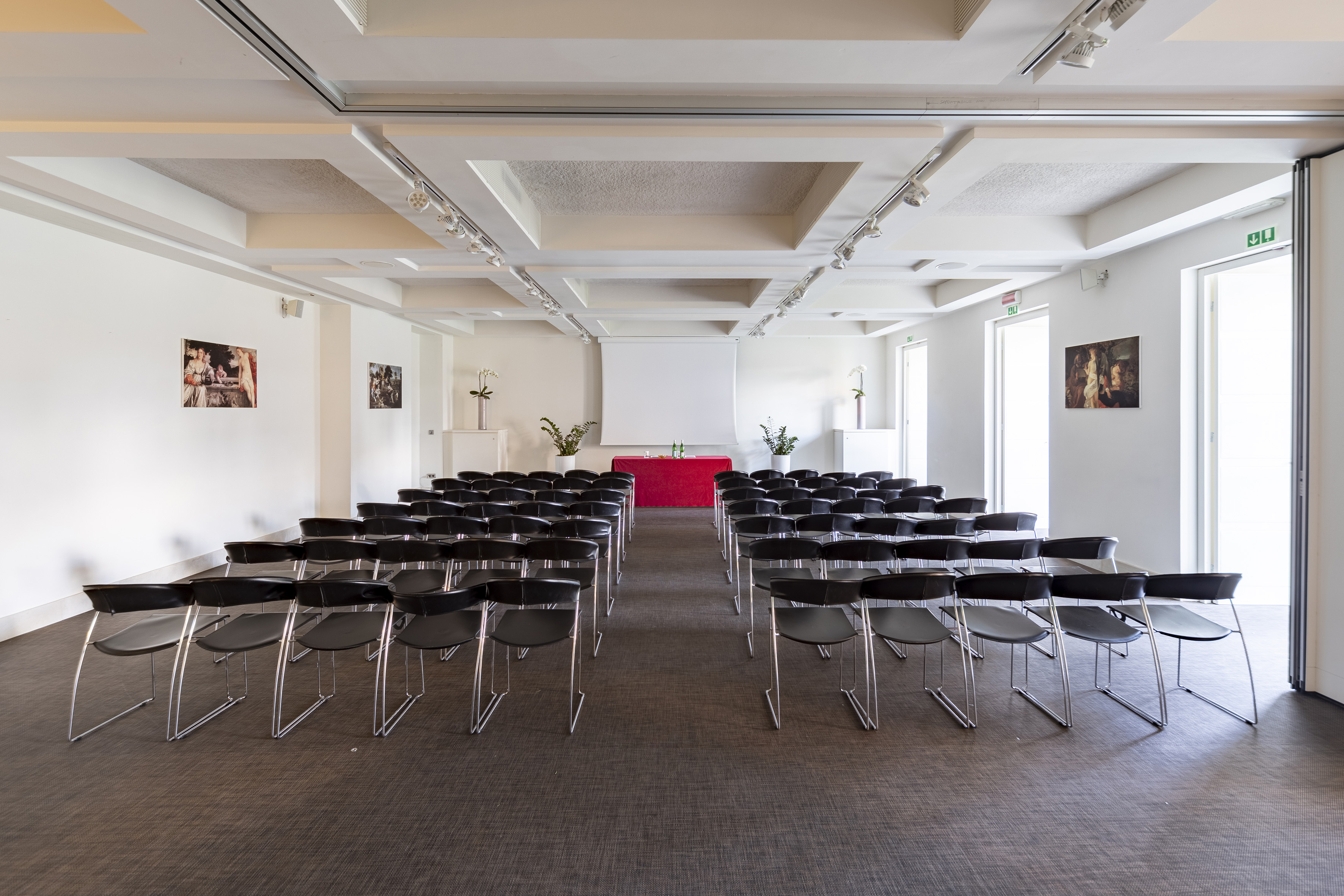 Modern meeting room with black chairs, projector screen, red table.