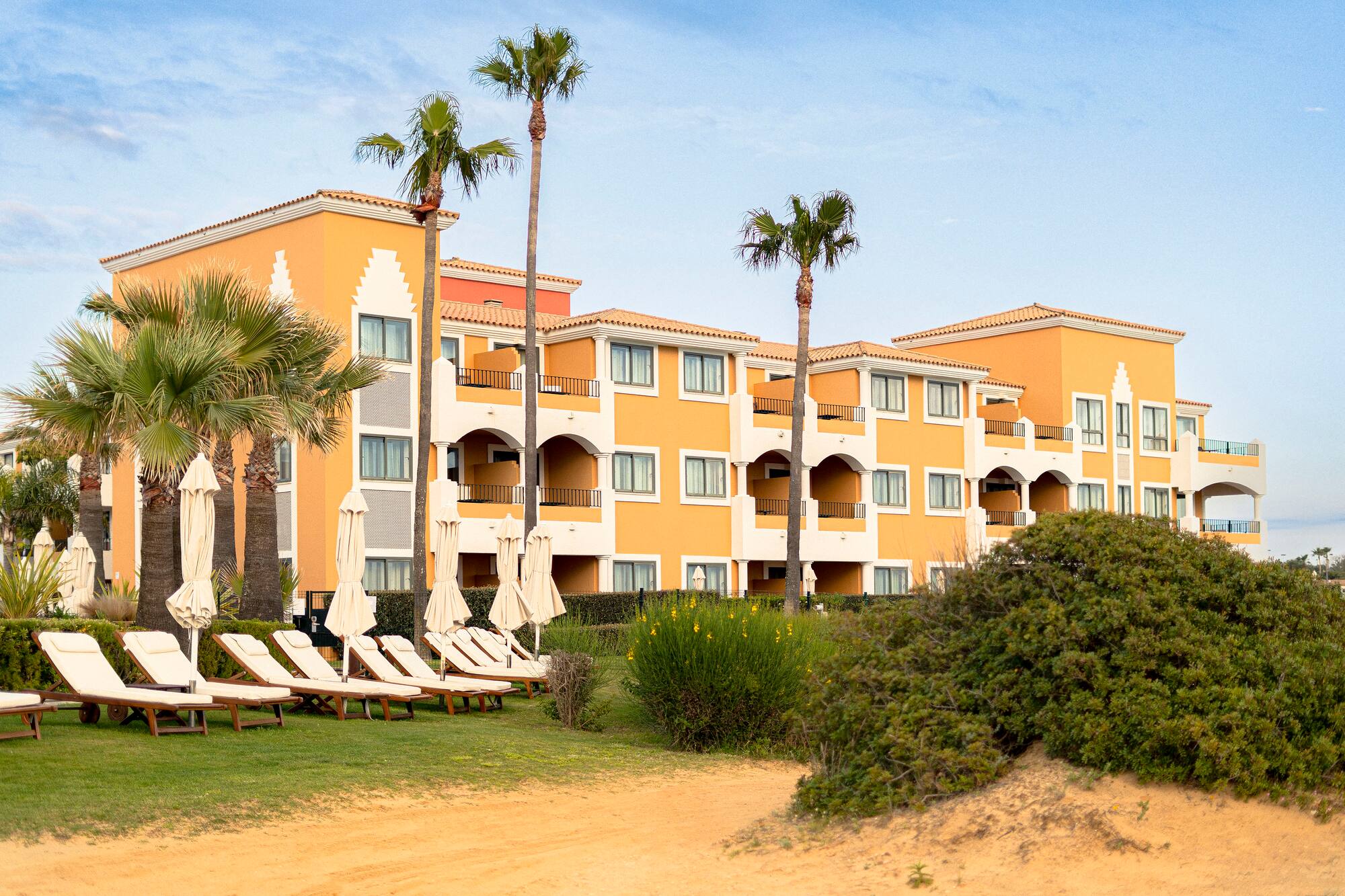 Orange beachfront hotel with palm trees, lounge chairs, and sand.