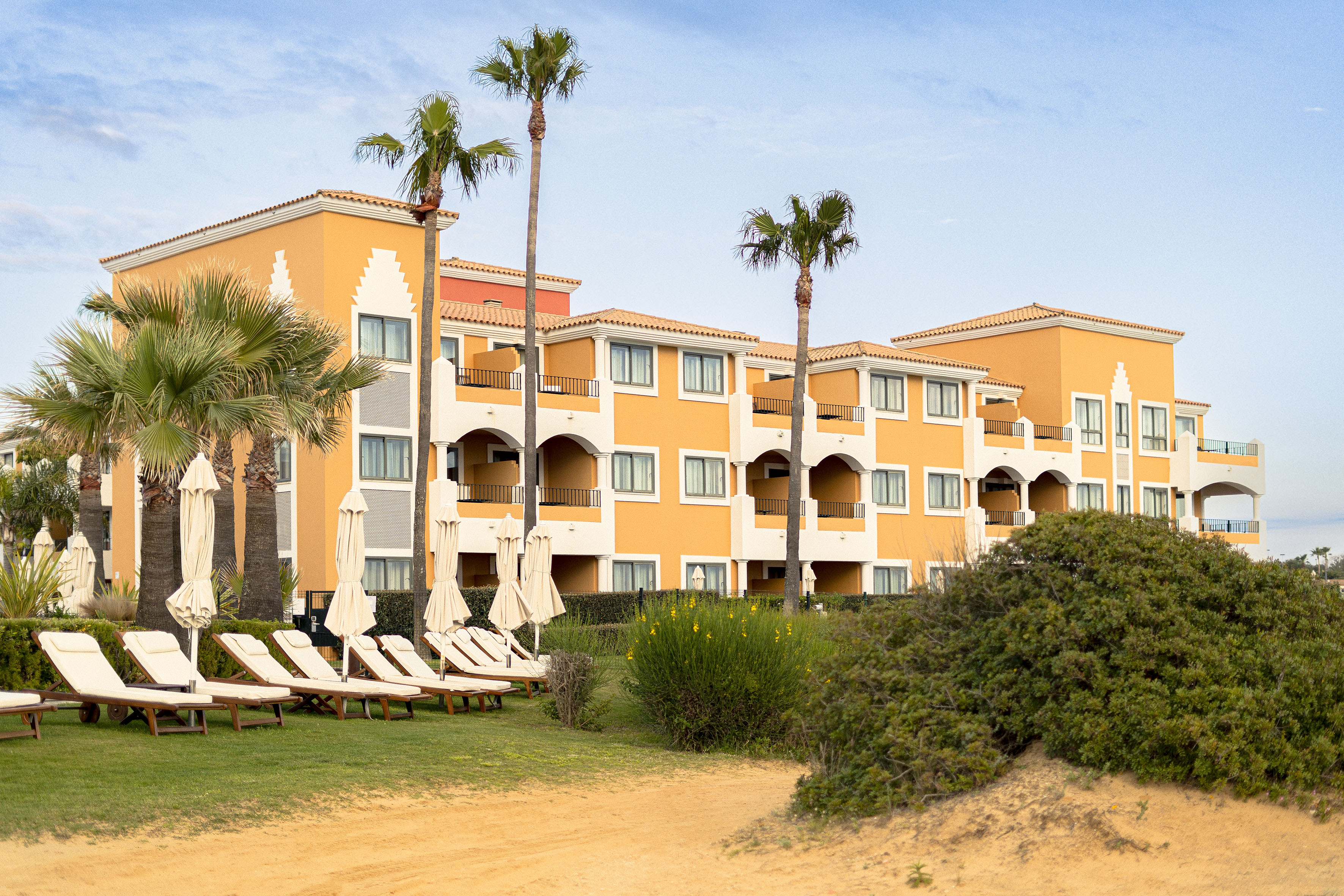 Orange beachfront hotel with palm trees, lounge chairs, and sand.