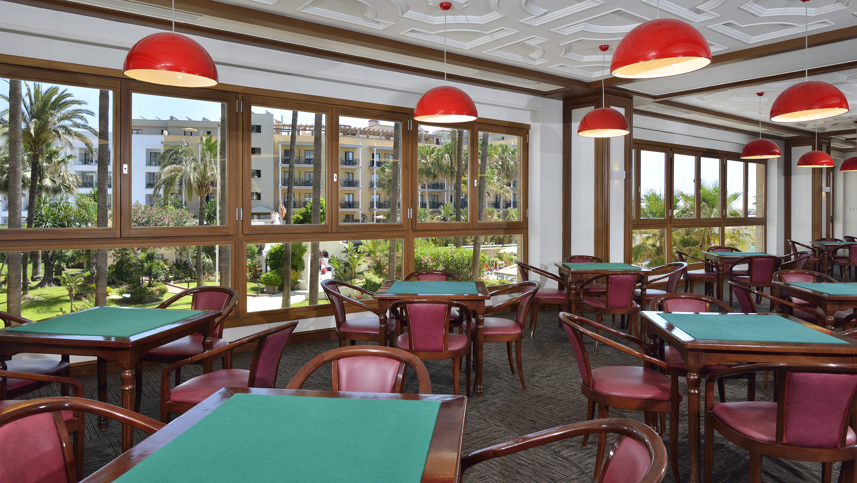 Hotel game room with green card tables, red chairs, and palm garden view.