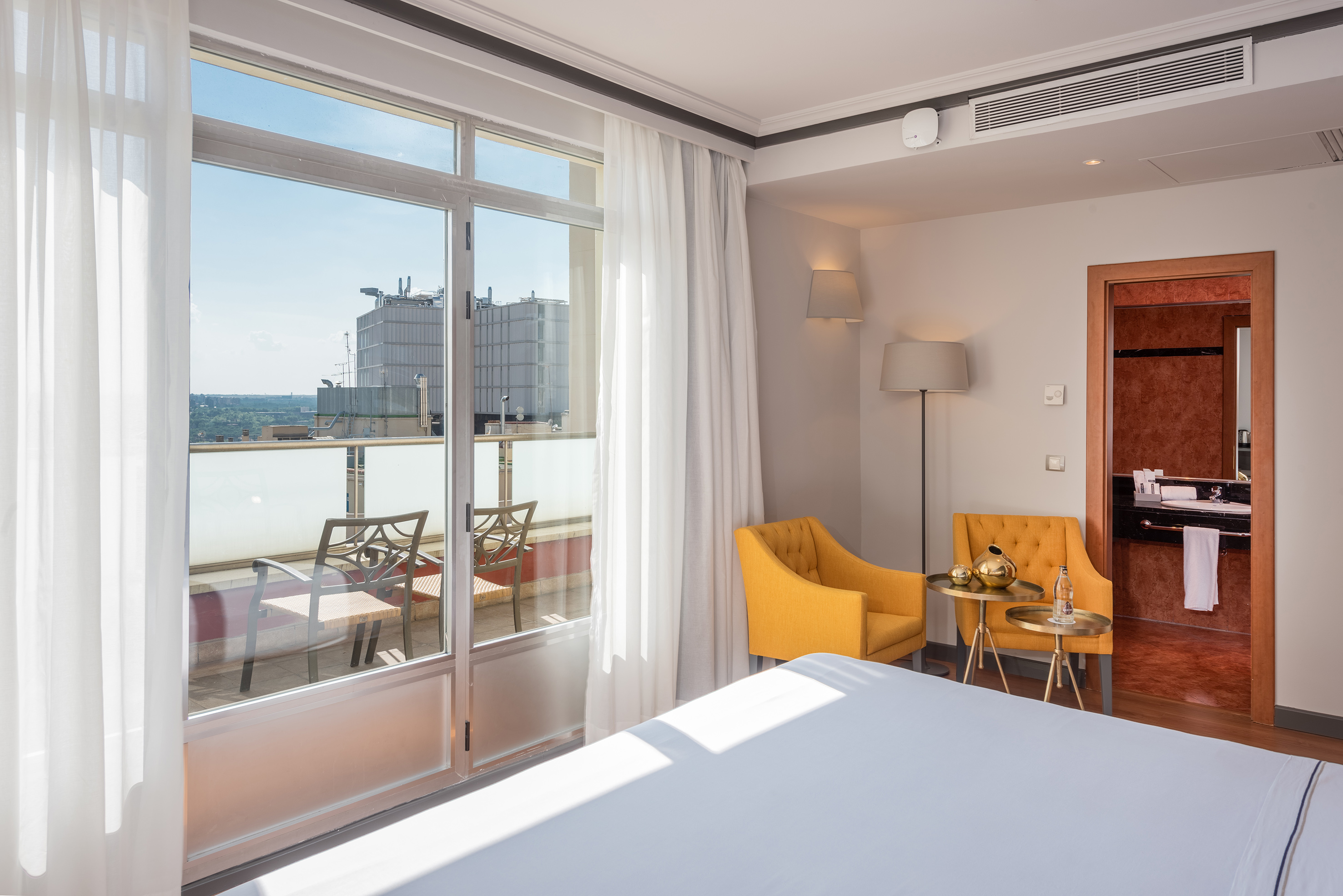Bright hotel room with balcony city view, yellow armchairs, and white bed.
